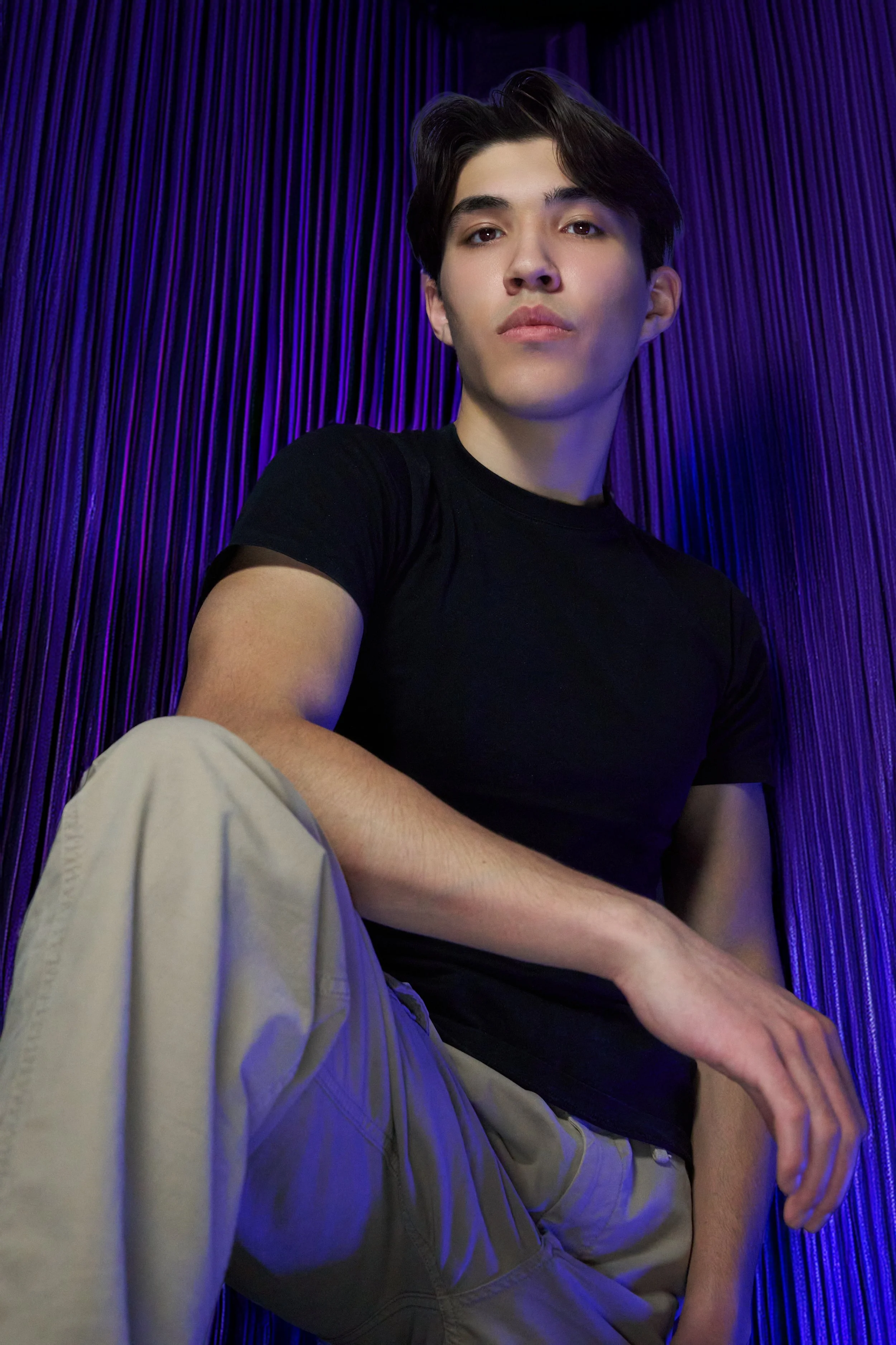 Young man with dark hair in black t-shirt and beige pants sitting against a purple curtain backdrop.