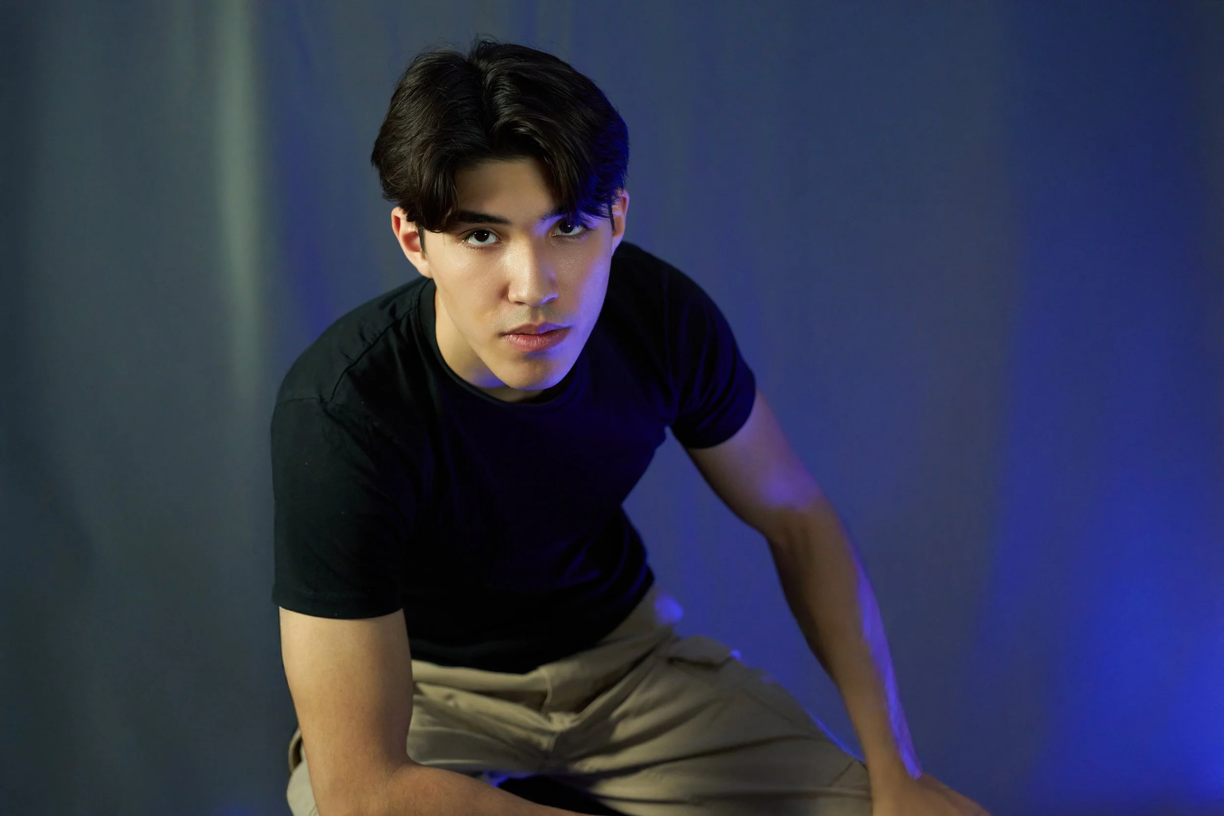 Young man with dark hair wearing a black T-shirt and beige pants, crouching and looking at camera against a dark background with blue lighting.