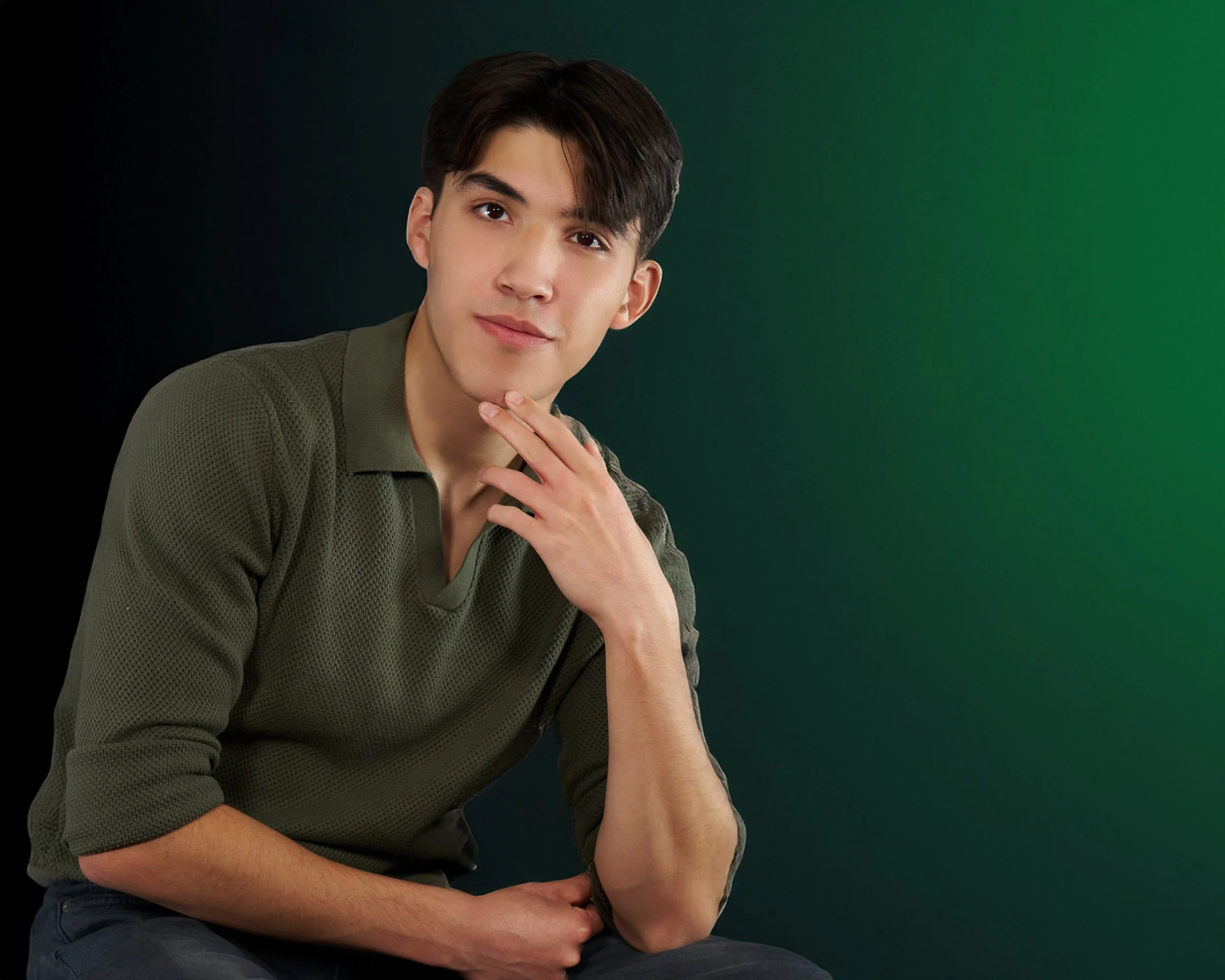 Portrait of a young man with dark hair, wearing a green textured long-sleeve shirt, posed against a gradient background transitioning from black to green.