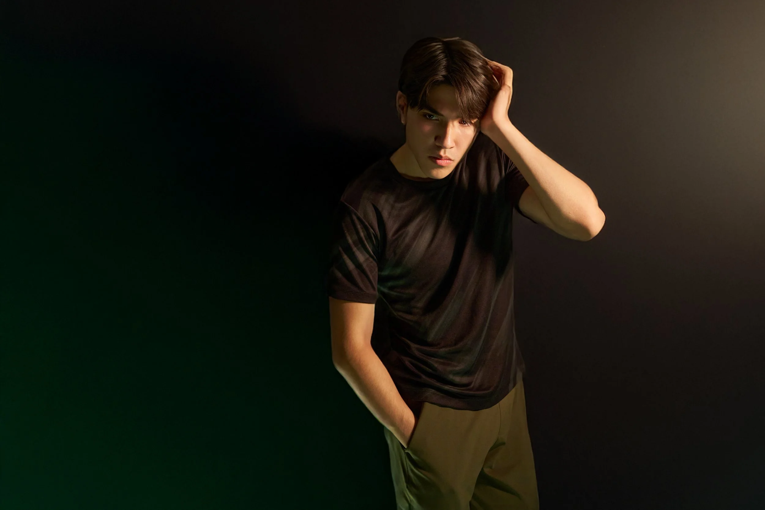 A young man with dark brown hair wearing a black t-shirt and beige pants, standing against a dark background with lighting on his face and body, looking at the camera with a serious expression, one hand touching his head.