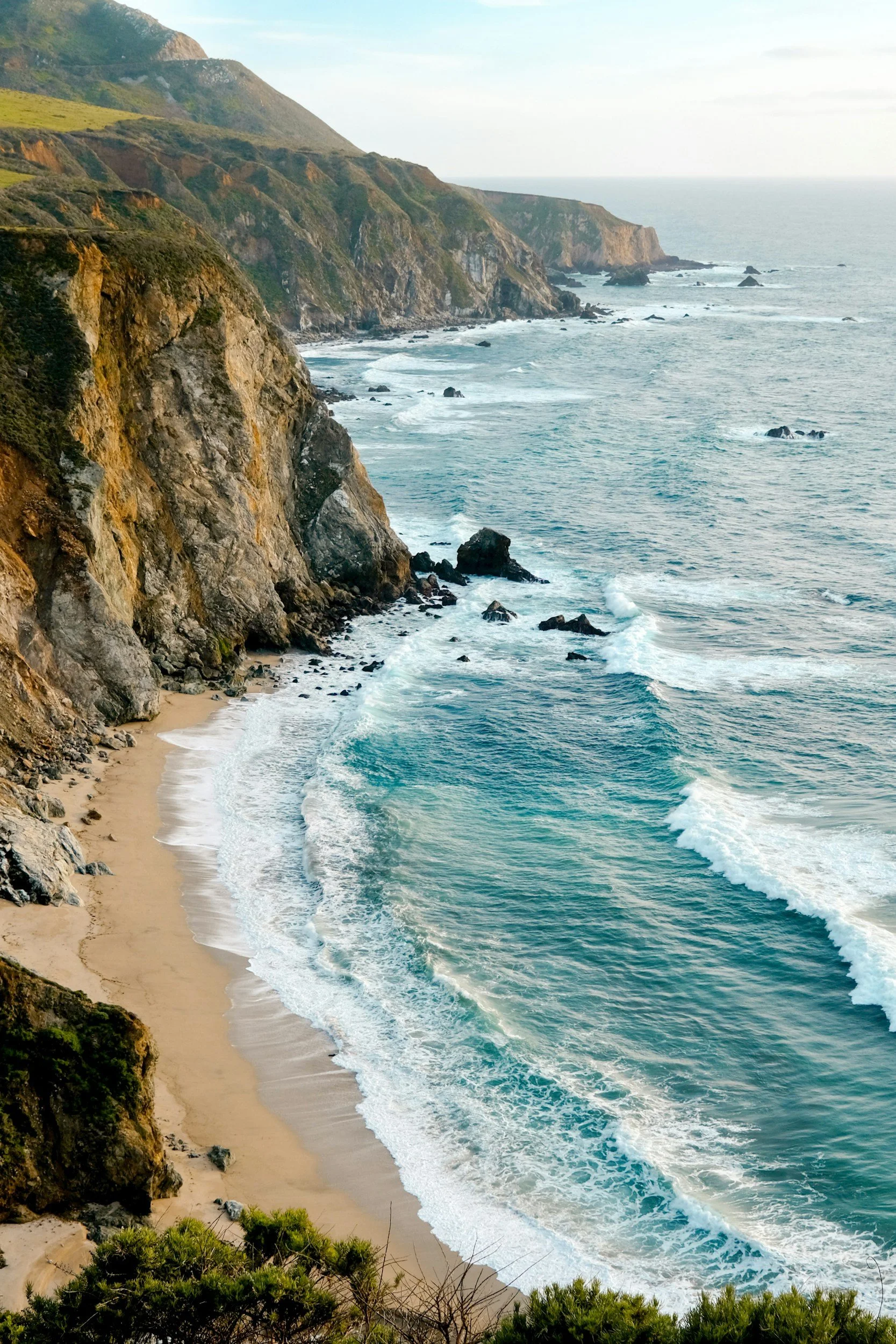 Coastal cliffs overlooking a sandy beach with waves crashing, under a partly cloudy sky.