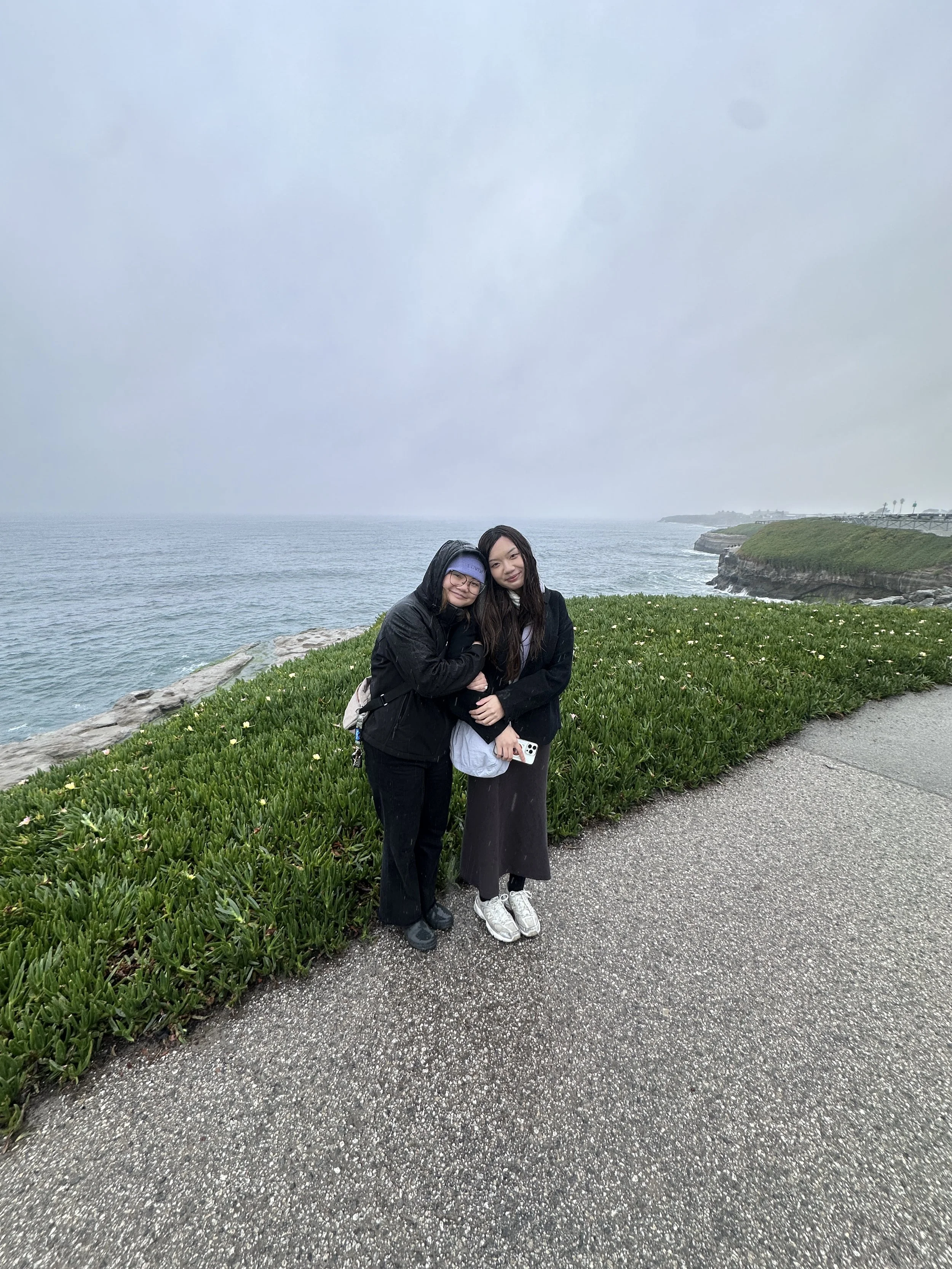 Two women standing together on a coastal pathway with the ocean and rocky shoreline in the background on a cloudy, rainy day.