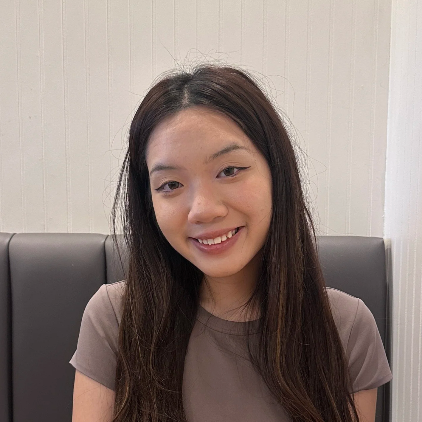 Kate, a young woman with long dark hair and tan shirt smiling while sitting on a booth.