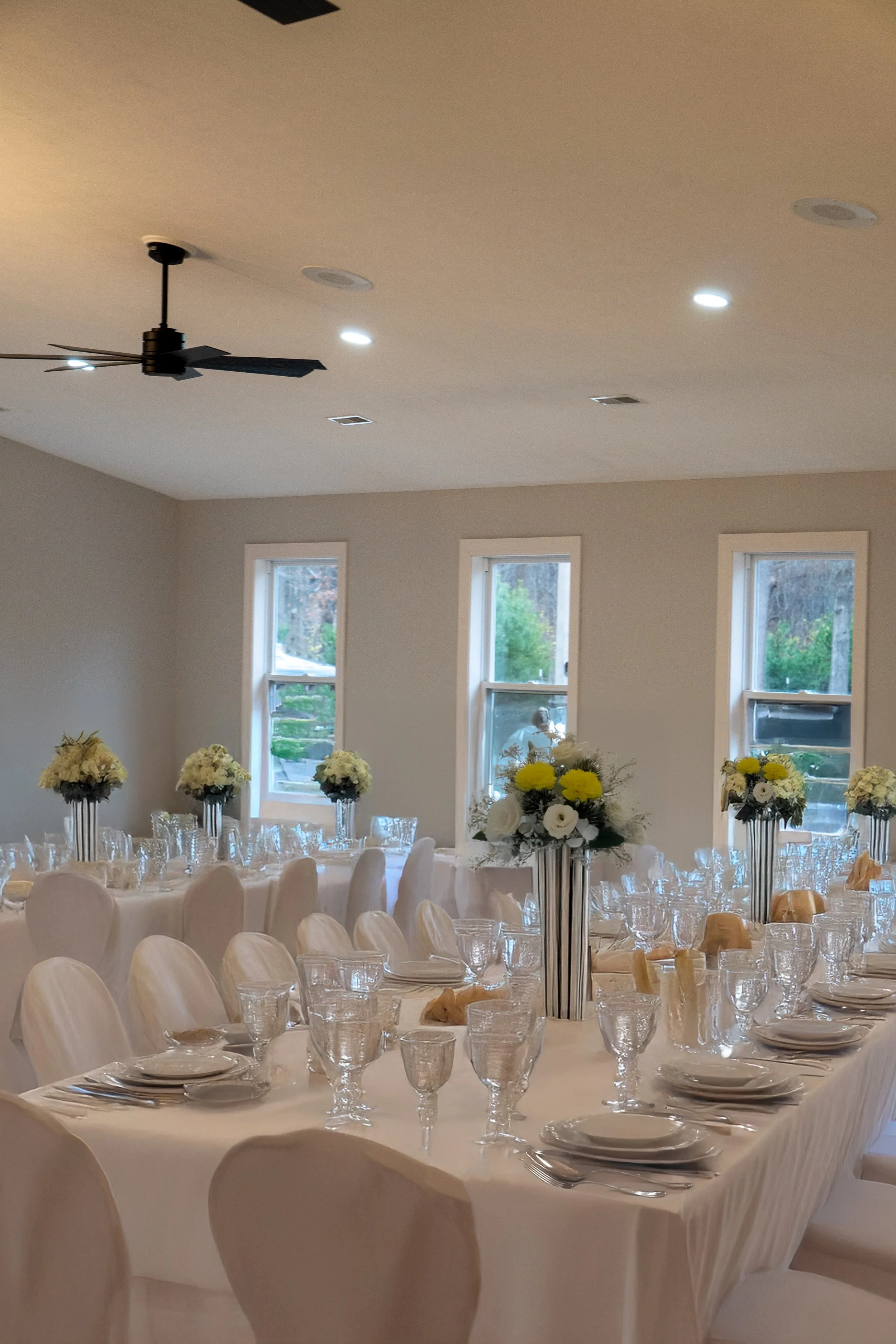 A banquet hall decorated for a formal event with round tables covered in white tablecloths, white chairs, glassware, and elegant floral centerpieces with white and yellow flowers.
