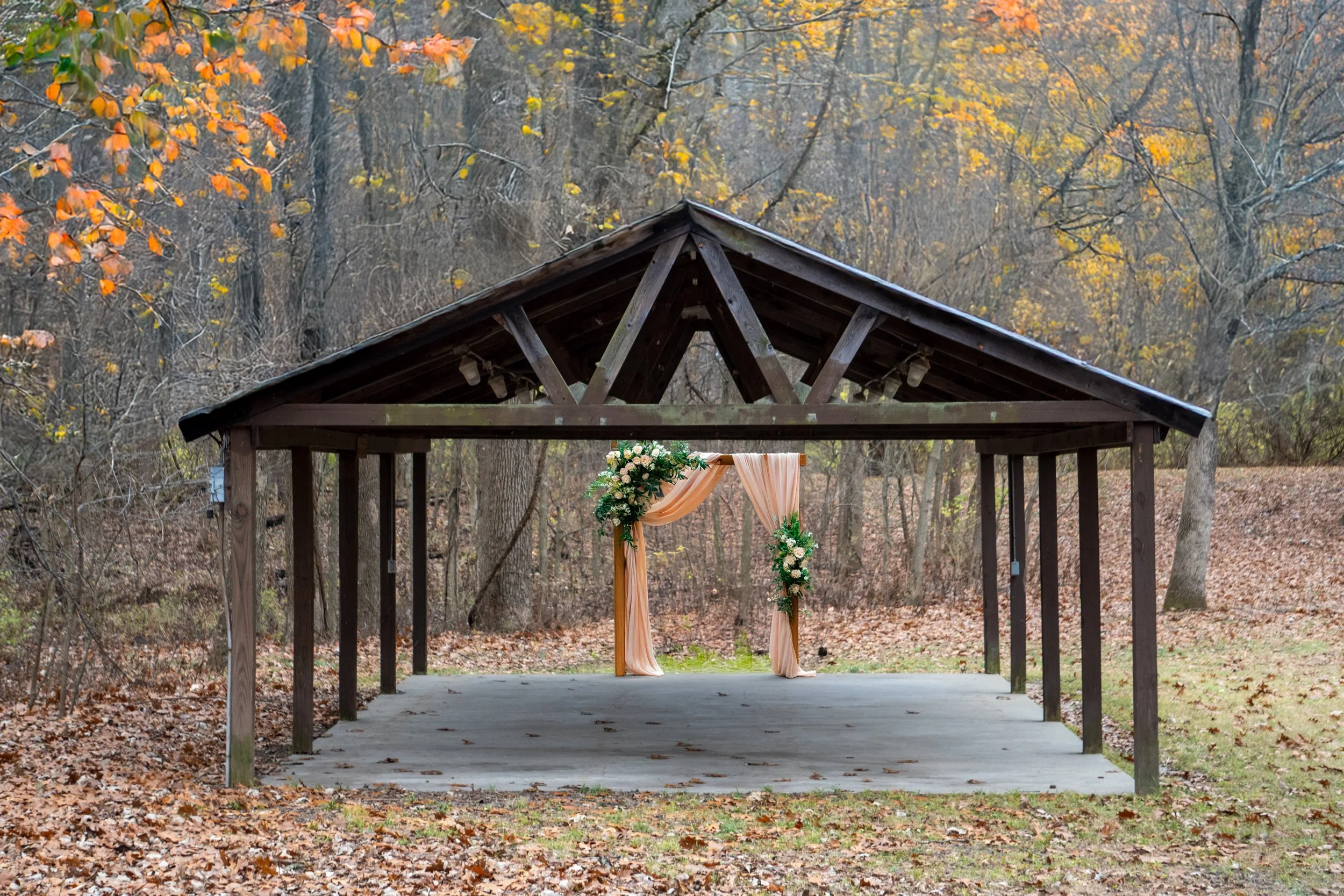 An outdoor pavilion decorated for a wedding ceremony with a floral and fabric arch, set amidst autumn trees with fall foliage.