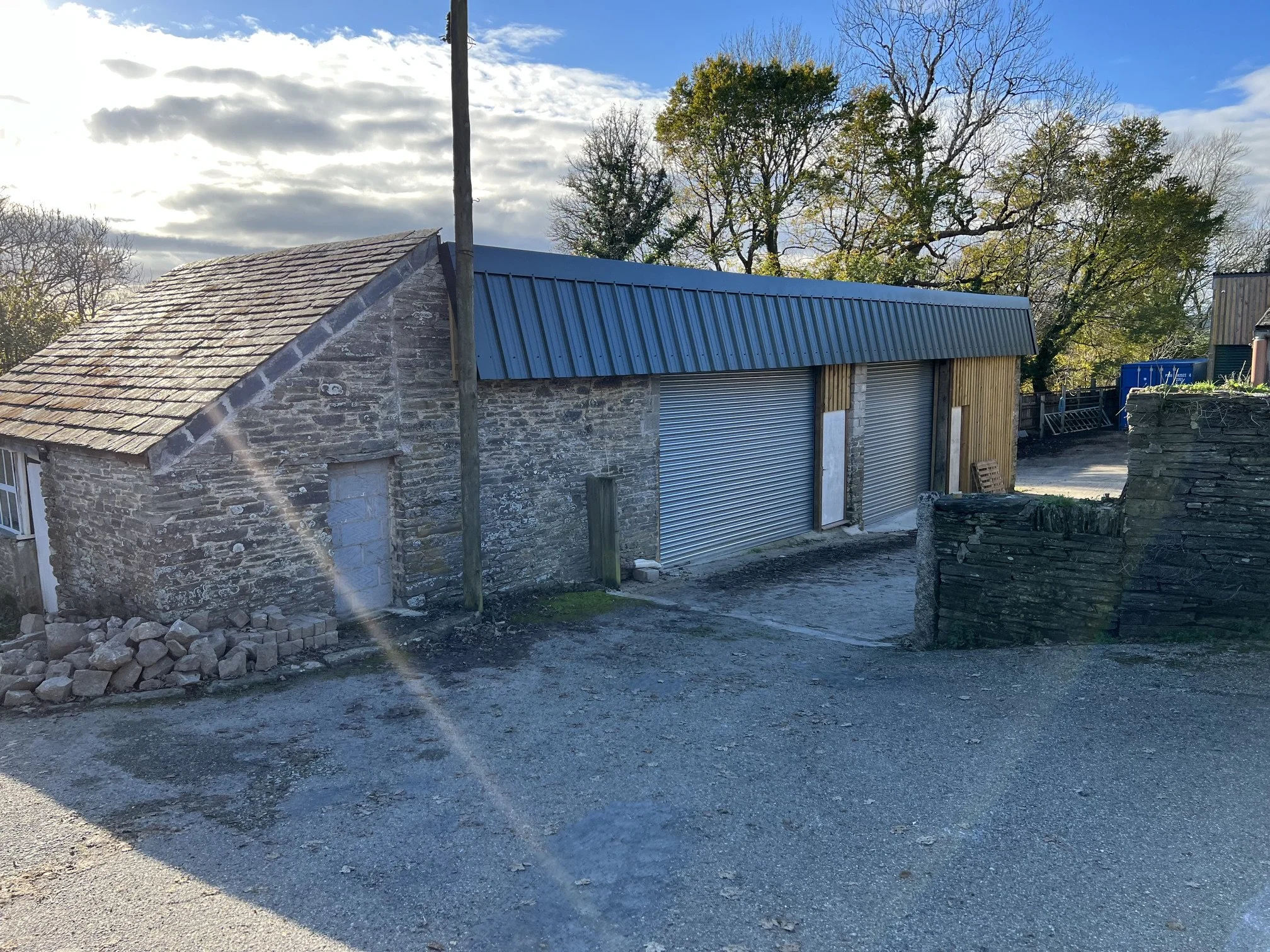 View of a rustic stone building with a metal roof and garage doors, surrounded by trees with some leaves and some bare branches, under a partly cloudy sky.