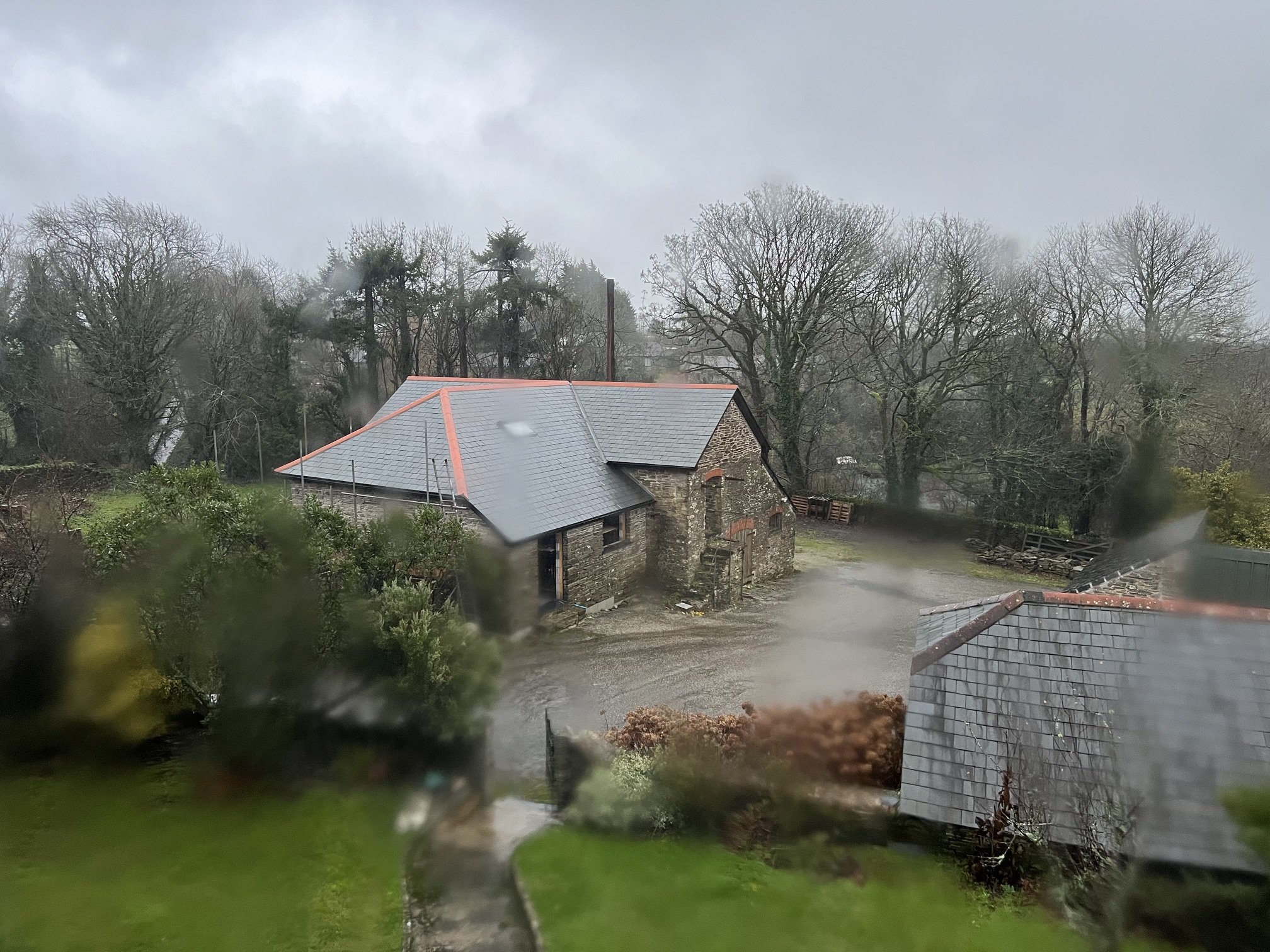 Rainy landscape showing a stone house with a grey roof, surrounded by leafless trees and wet ground, viewed through a rain-spattered window.