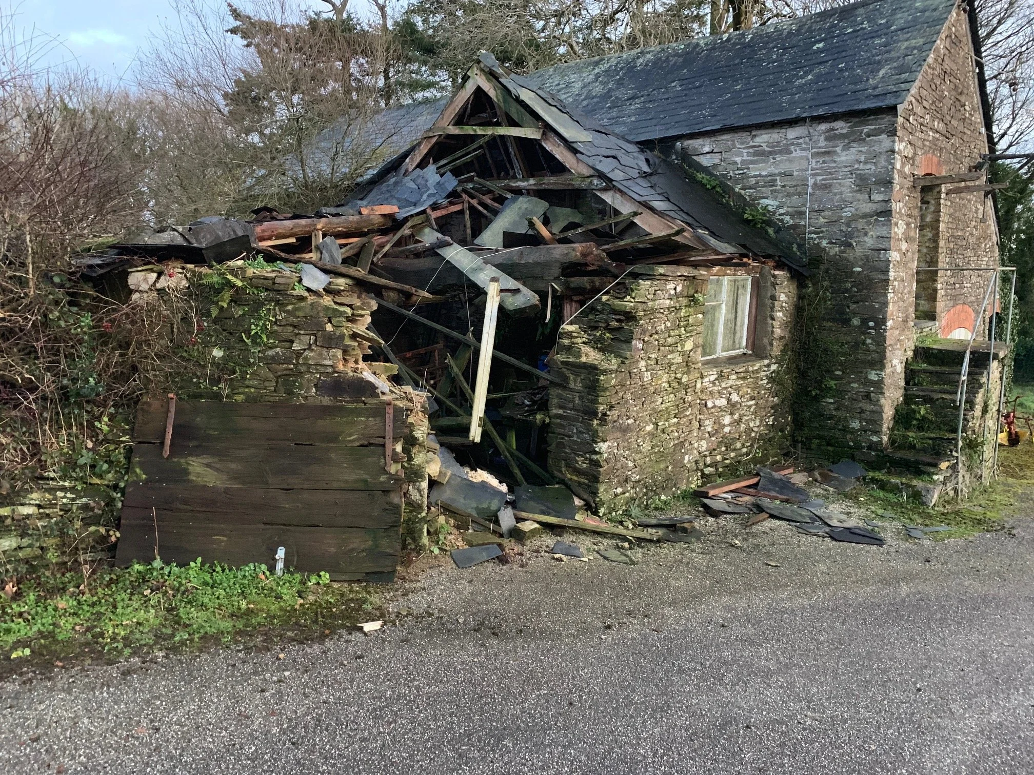 A damaged stone and wood house with part of the roof collapsed showing wooden beams and debris, surrounded by overgrown bushes and trees, next to a gravel pathway.