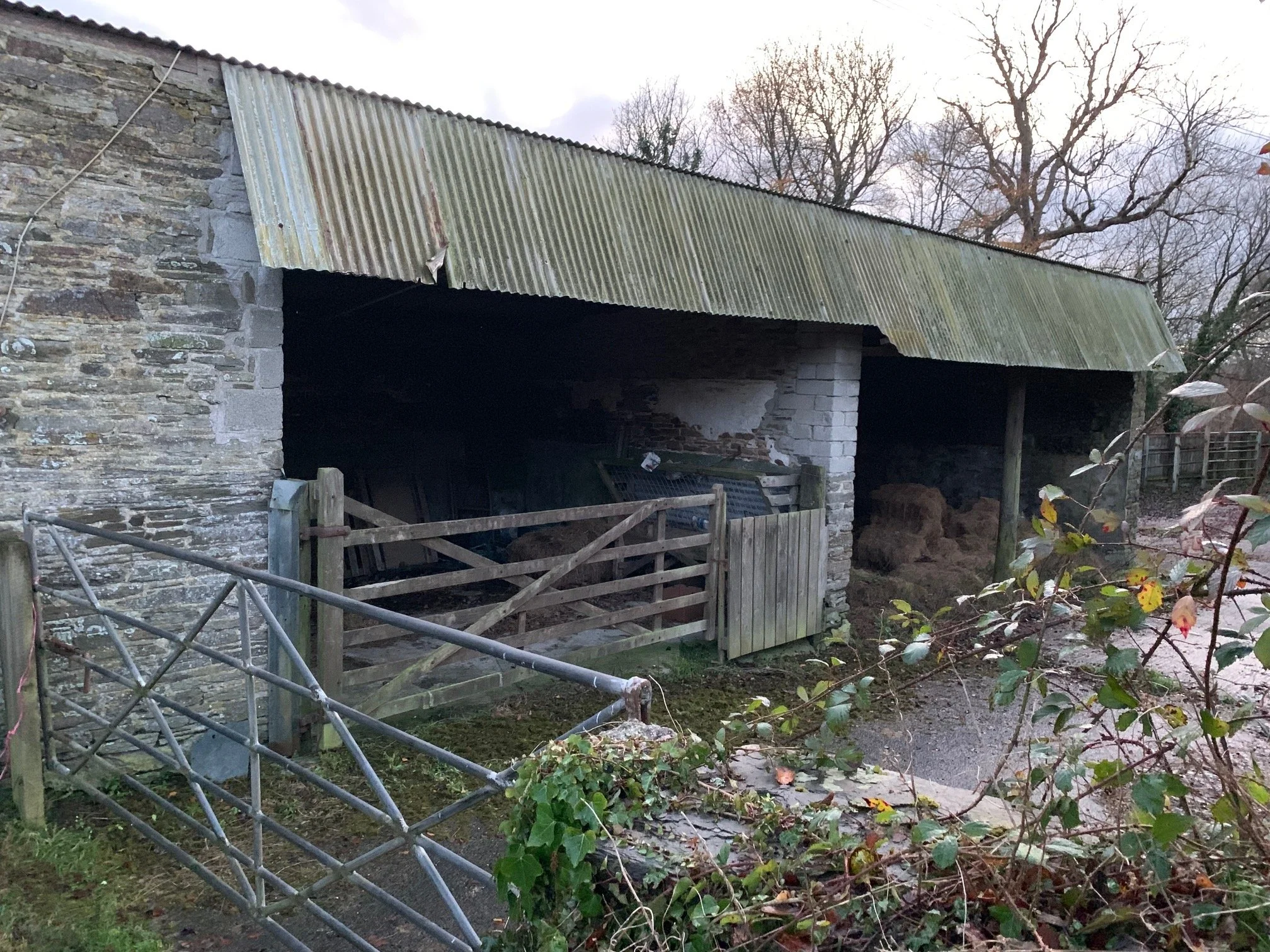 Old stone and brick barn with a weathered corrugated metal roof surrounded by a wooden fence and overgrown trees with autumn leaves.