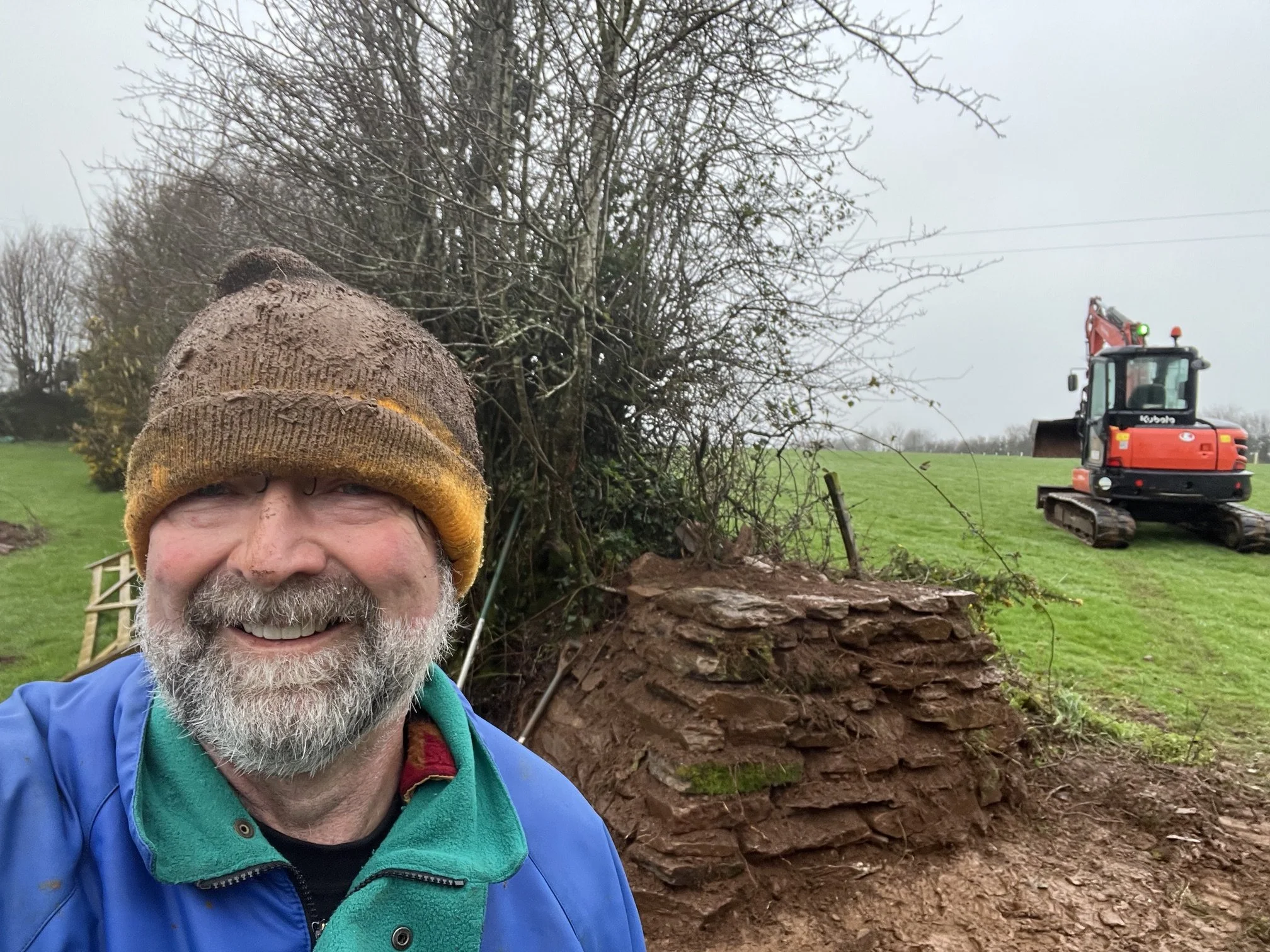 A man with a white beard wearing a yellow knit cap and a blue jacket takes a selfie in front of a fallen tree while doing outdoor work with a small orange and black excavator in the background on a cloudy day.