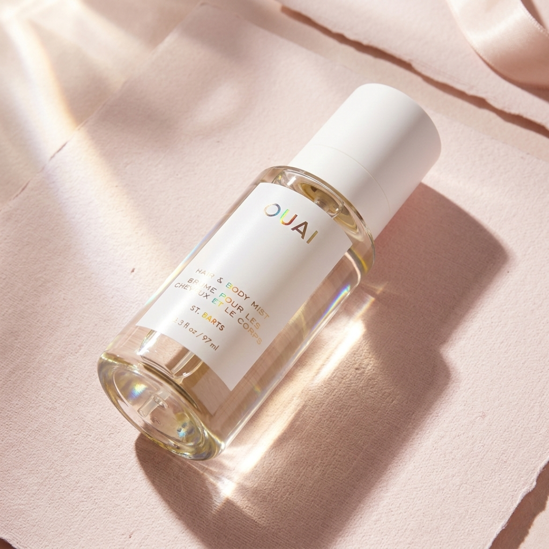 A minimalist bottle of OUAI St. Barts Hair & Body Mist with a white cap, lying diagonally on layered sheets of pale pink textured paper. Shimmering light reflects off the clear liquid and the holographic lettering on the label.