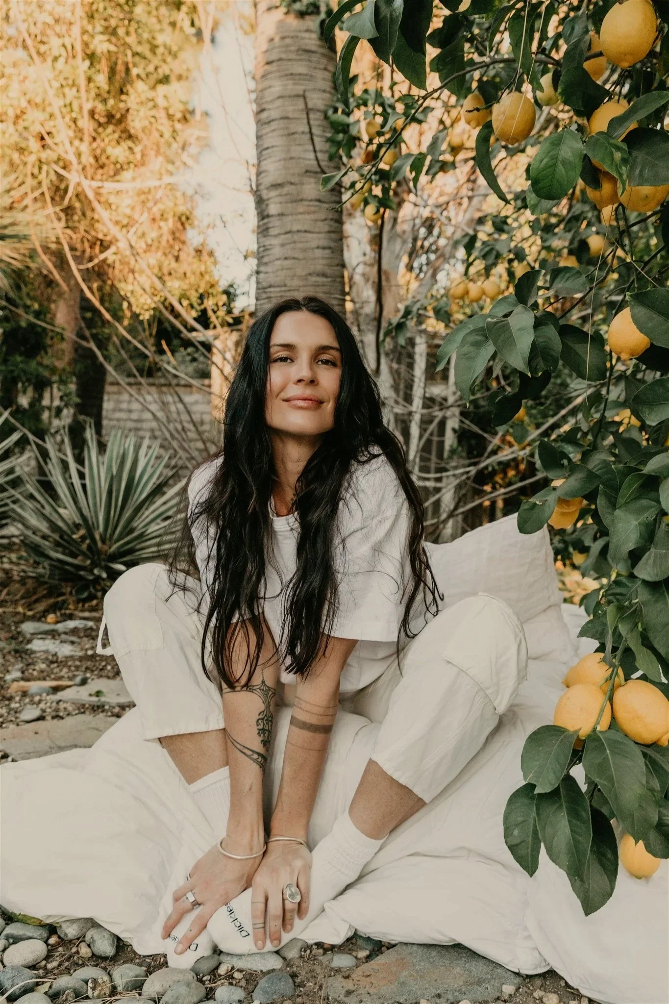 A young woman with long dark hair, tattoos, and jewelry sitting outdoors among trees and yellow fruit hanging from a branch, wearing a white outfit with a relaxed expression.