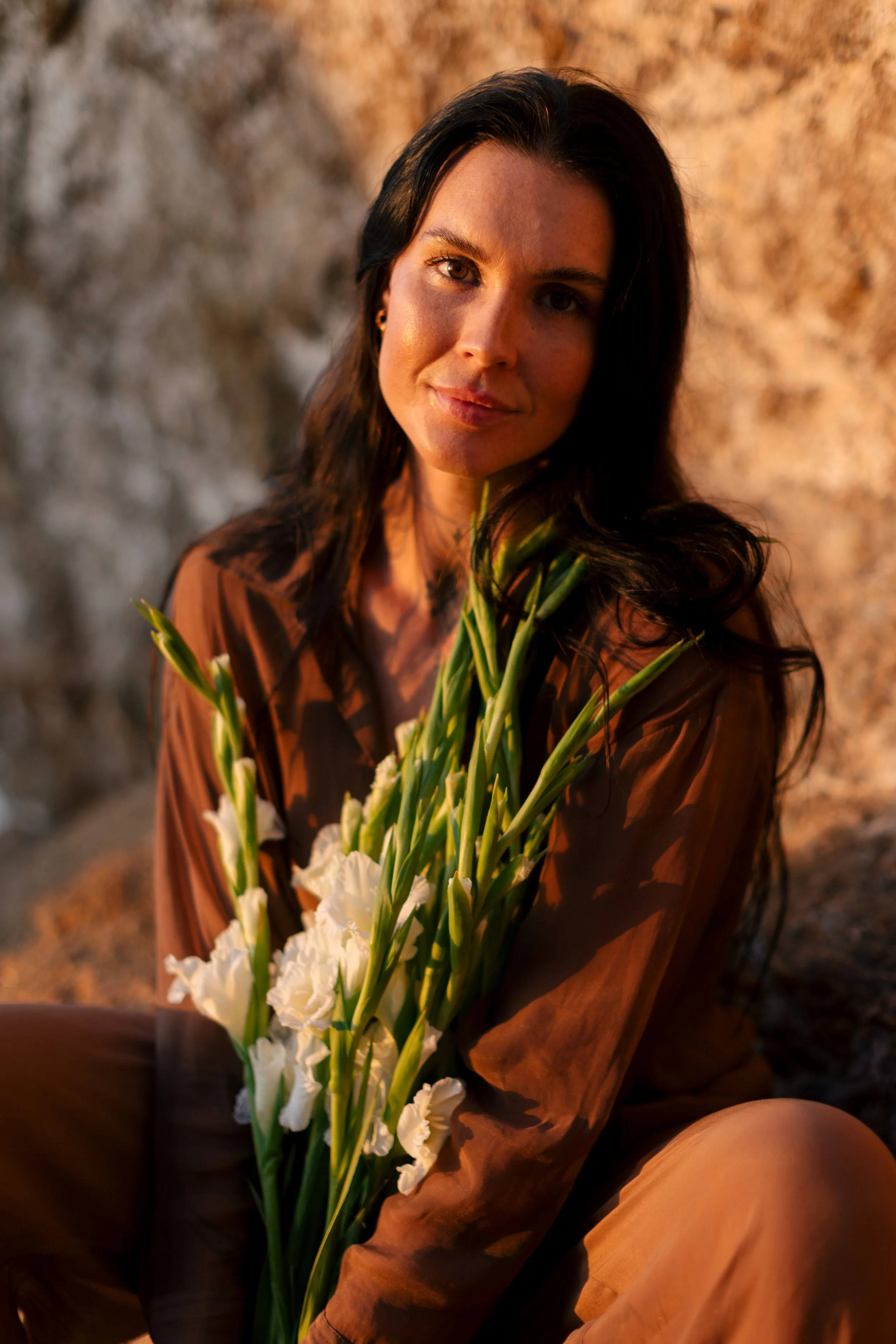 A woman with long dark hair holds a bouquet of white flowers, sitting against a rocky background, bathed in warm, natural sunlight.