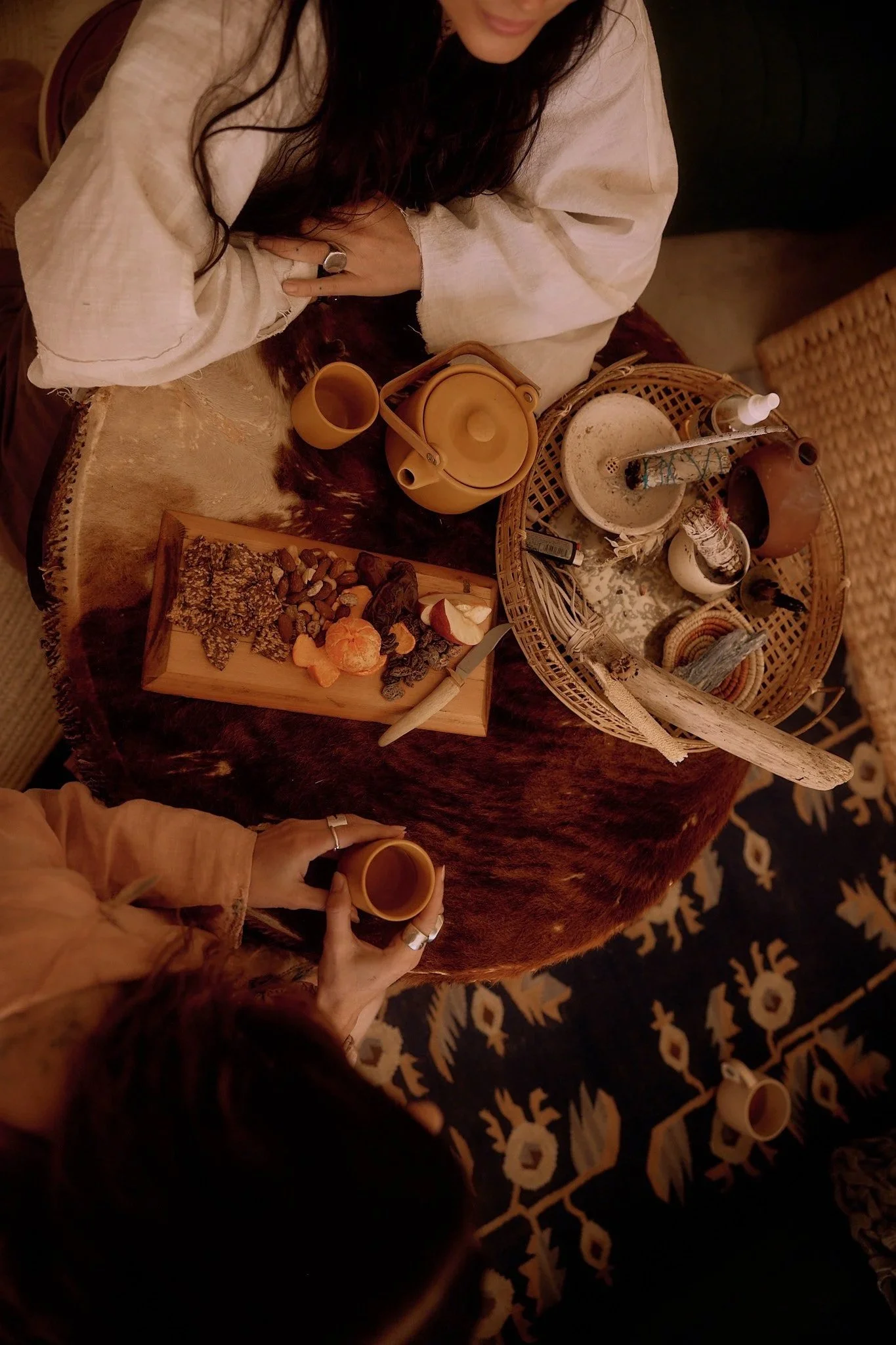 A woman with long dark hair wearing a cream-colored top sitting at a table with various tea utensils, a wooden tray with snacks, and a person holding two small cups with tea. The setting appears cozy and warm with ethnic patterned rugs visible.