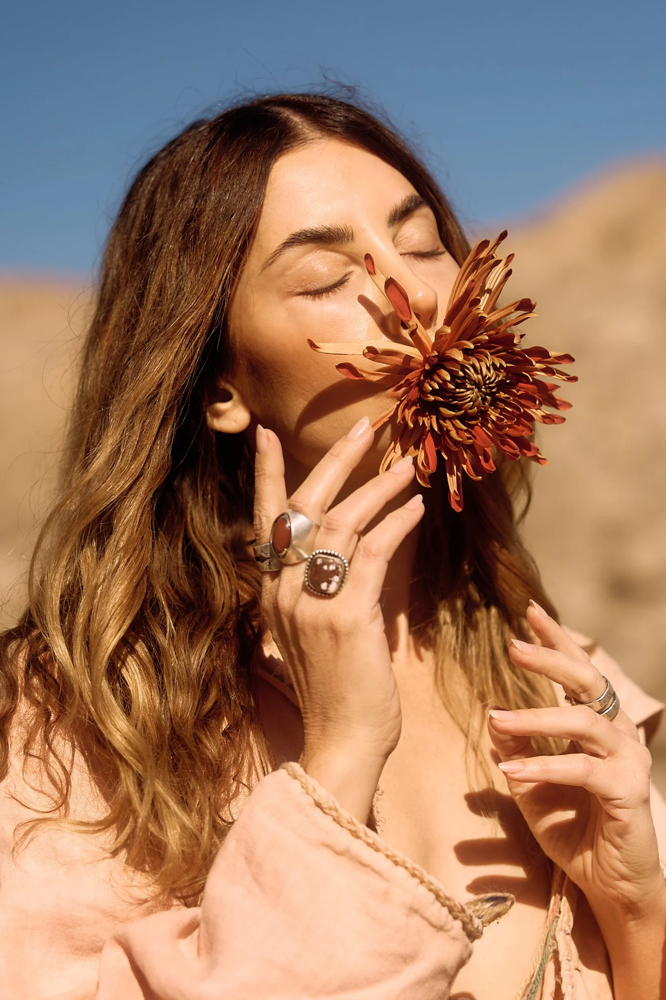A woman with long, wavy brown hair and jewelry holds a large brown-orange flower close to her face with her eyes closed, outdoors in sunlight.