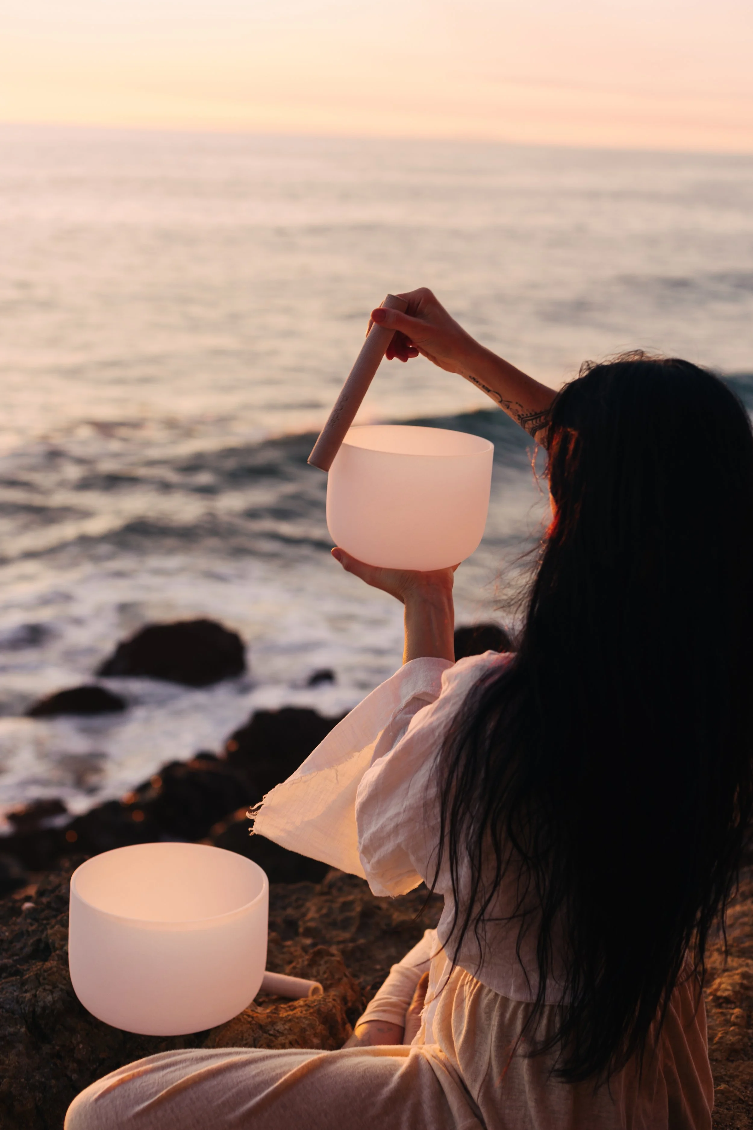 A woman sitting on rocks at the beach holding a singing bowl in one hand and a mallet in the other, with the ocean and a sunset in the background.