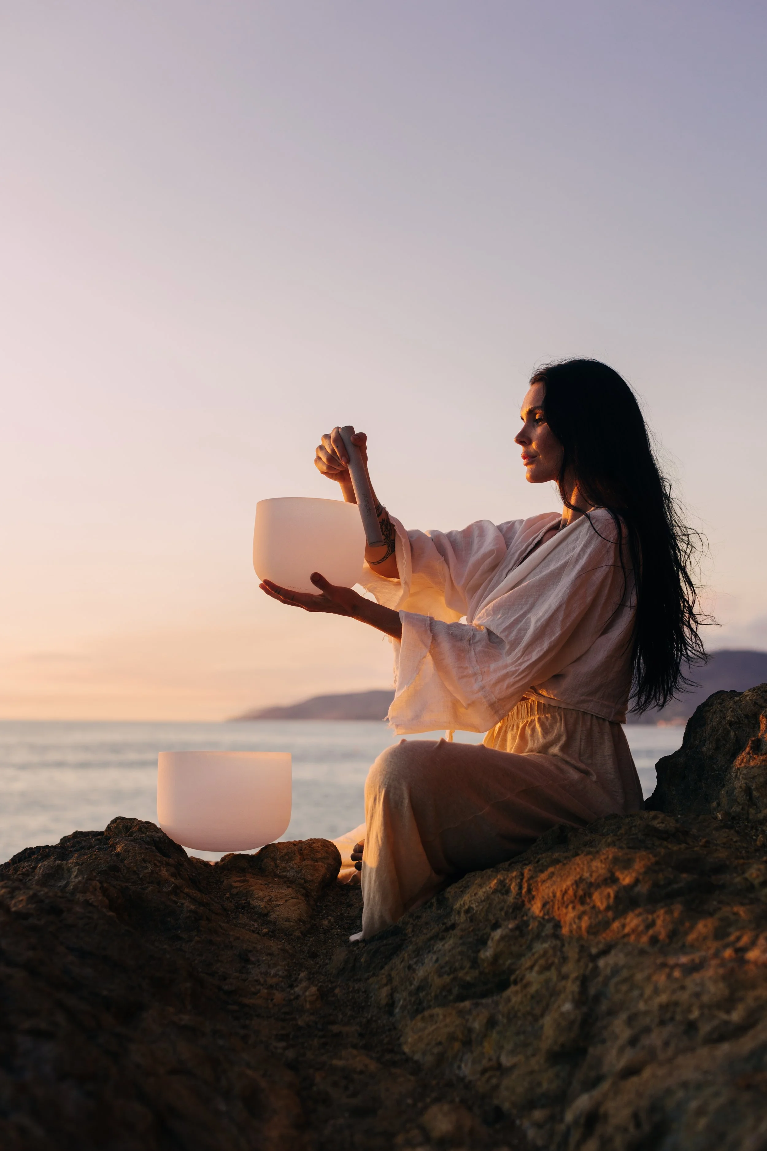 A woman sitting on rocks by the ocean at sunset holding a singing bowl, with a candle nearby and calm waves in the background.