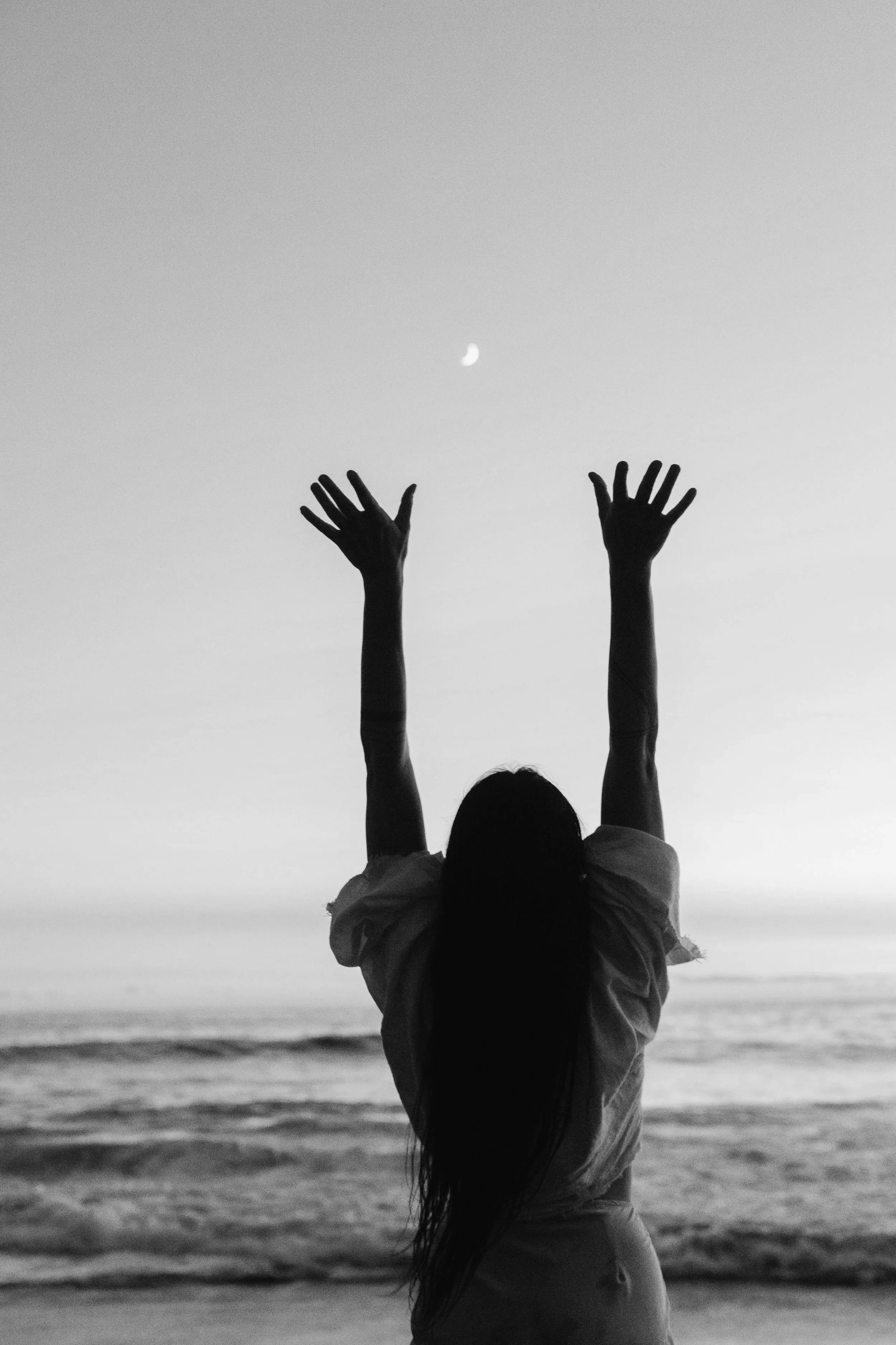 Silhouette of a girl with long hair standing on the beach at sunset, reaching up towards the moon in the sky.