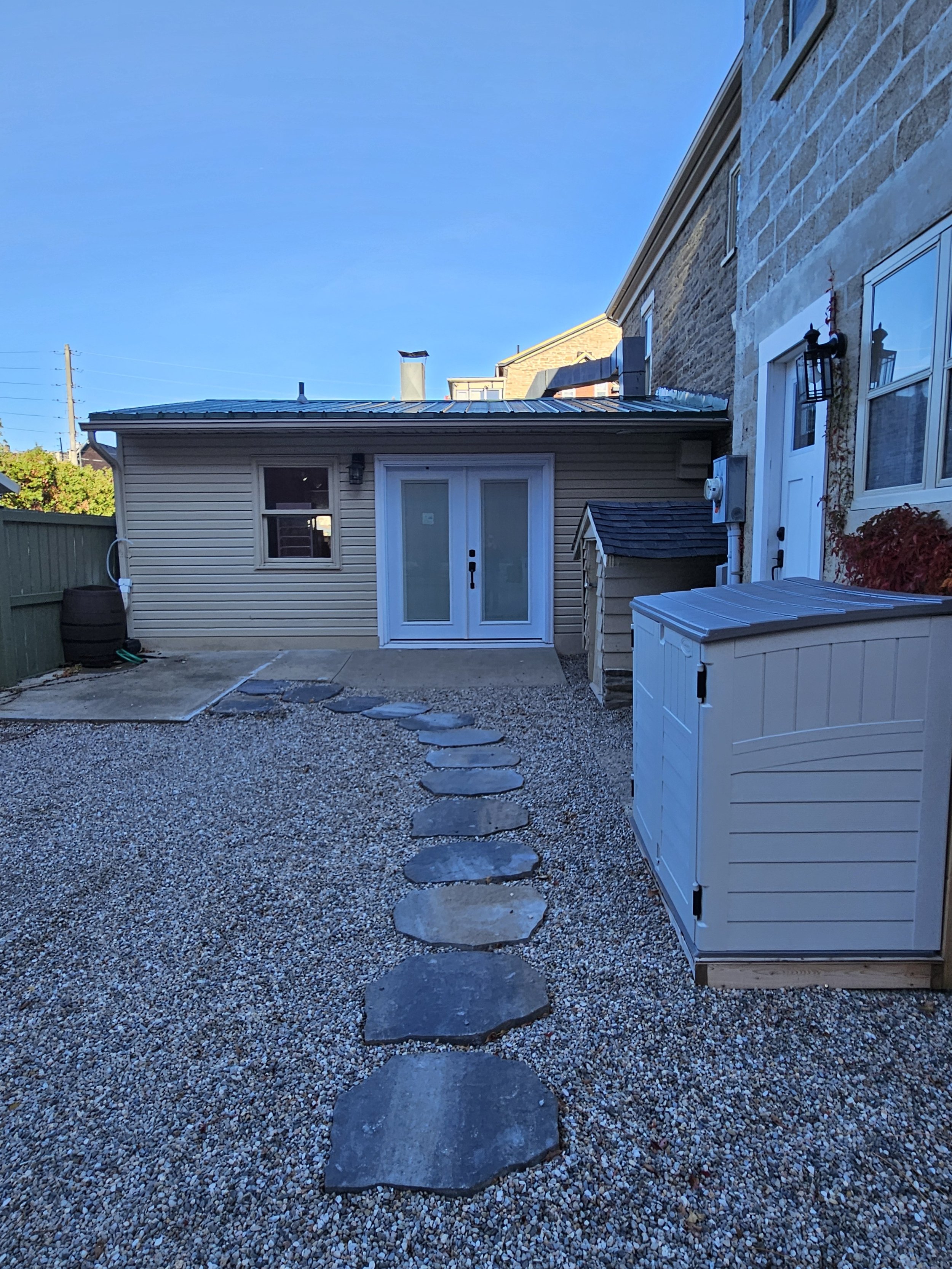 Gravel courtyard with stepping stones, a detached garage, stone house walls, and a large outdoor storage box under a clear blue sky