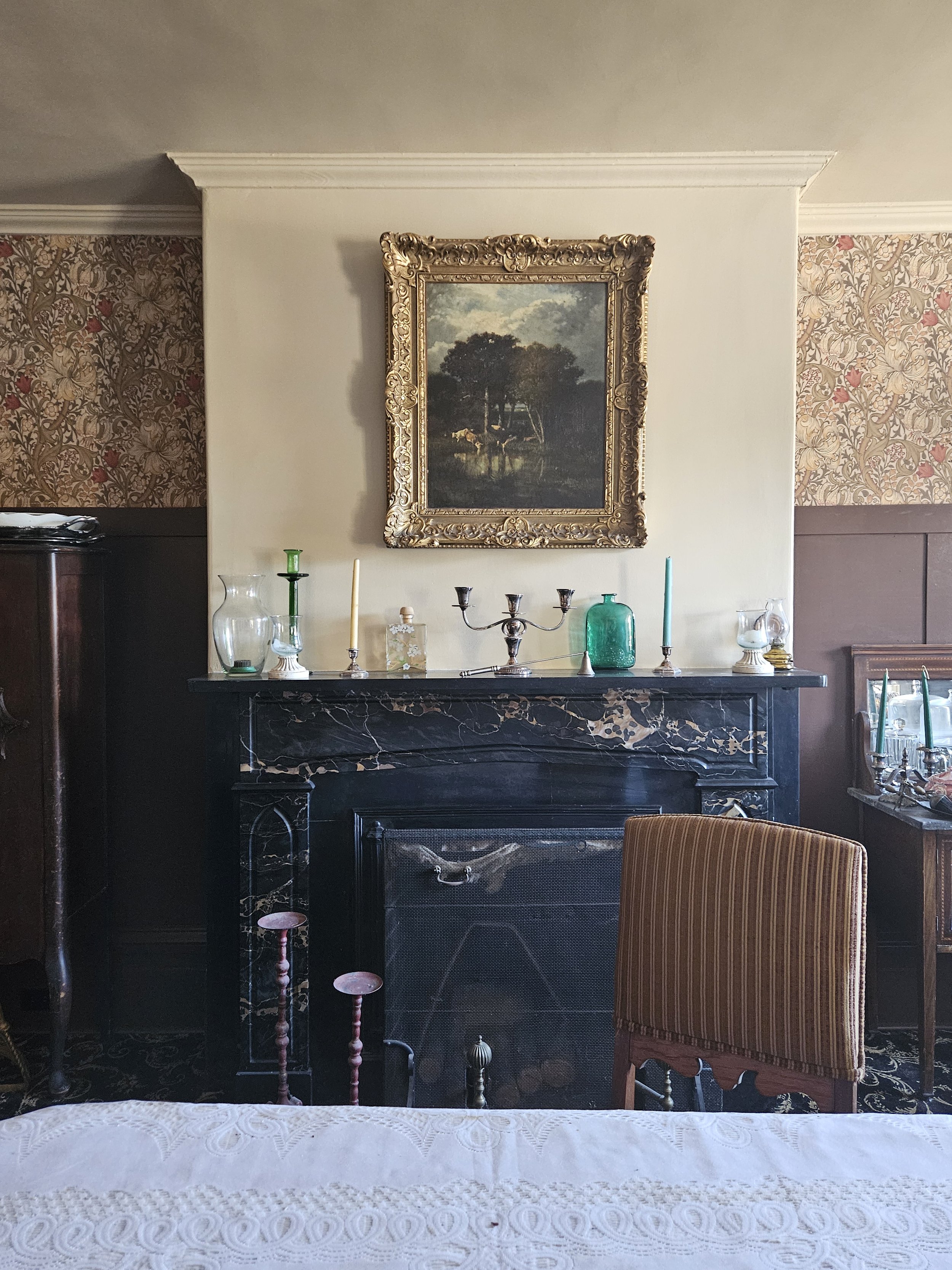Elegant room with a black marble fireplace, ornate framed landscape painting, floral wallpaper, candles, glass bottles, and a striped armchair