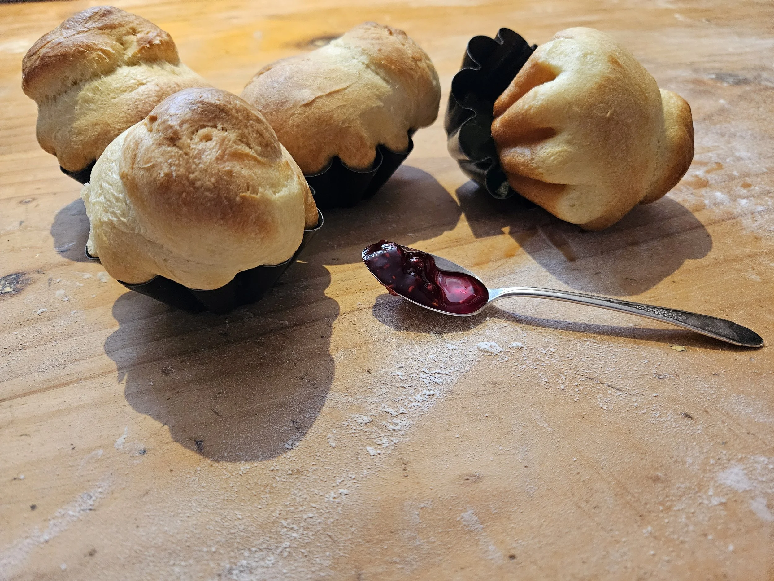 Fresh baked bread rolls in black baking cups with a spoonful of berry jam on a wooden table
