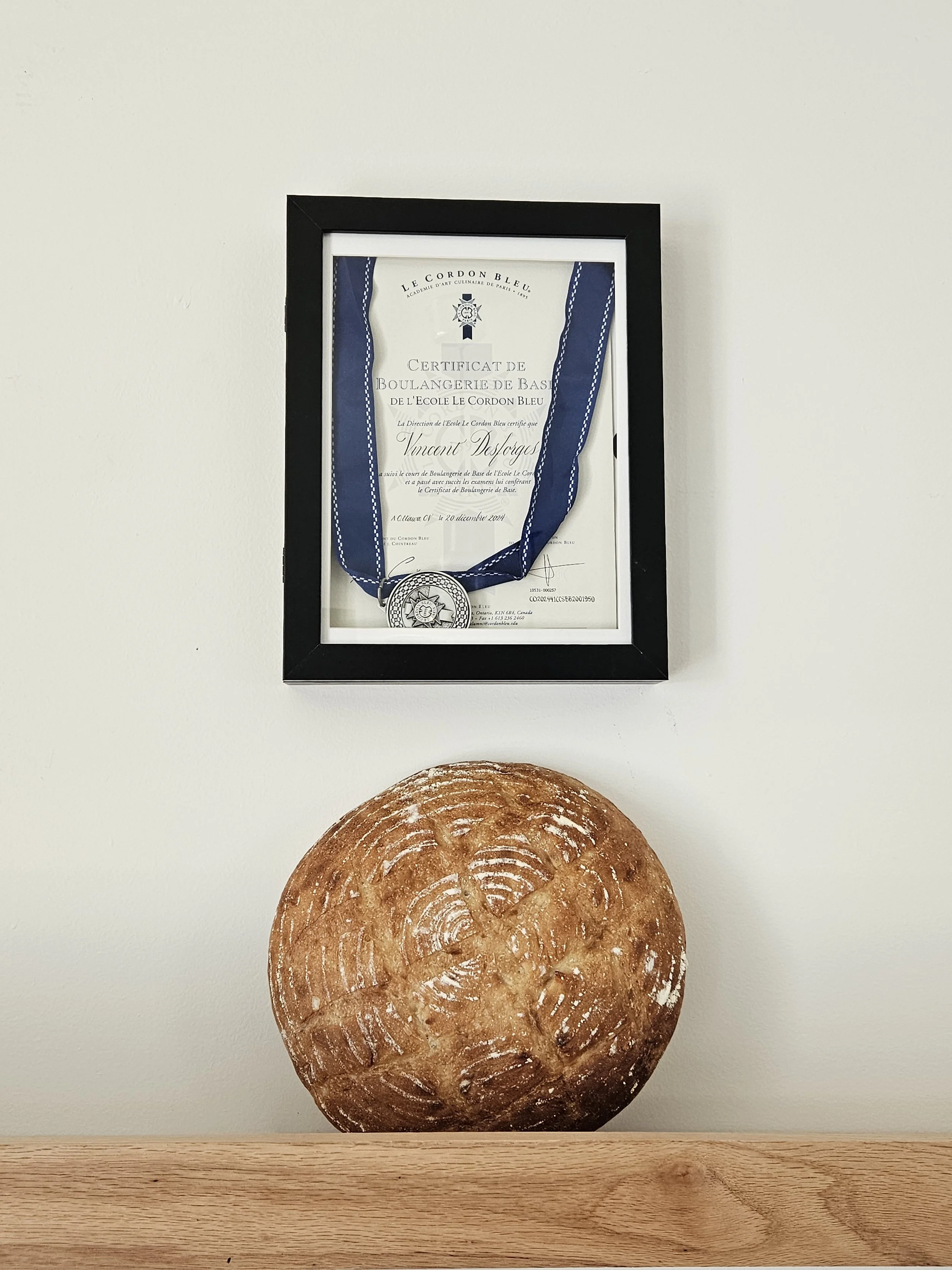 Framed Le Cordon Bleu basic baking certificate with a medal hanging over it, displayed above a rustic round loaf of bread.