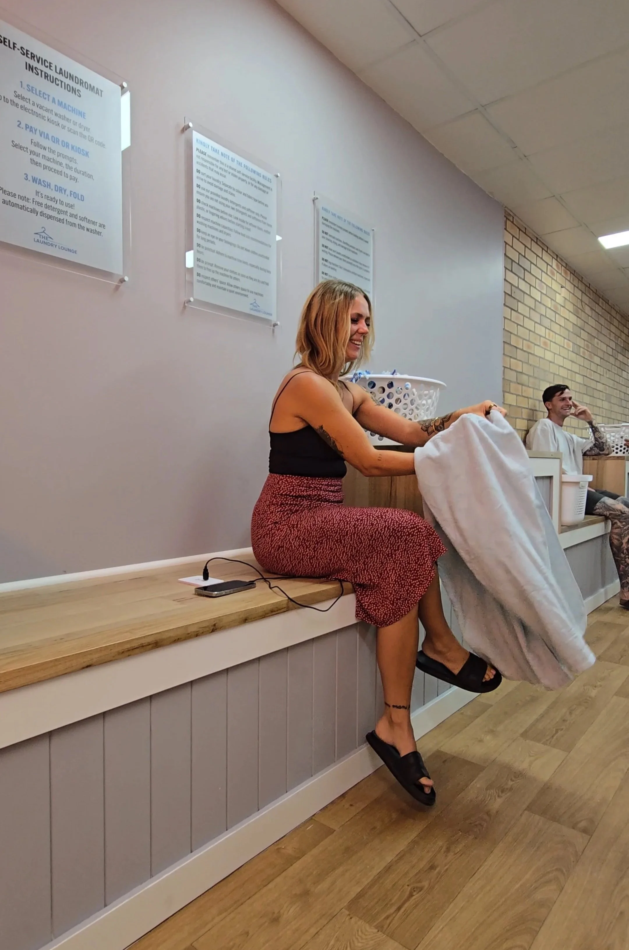 A blonde woman in a black top and red skirt sits on a bench seat. She smiles while folding a towel, next to her her phone is on charge from a complementary charging port. In the background a young man smiles while waiting for his machine.