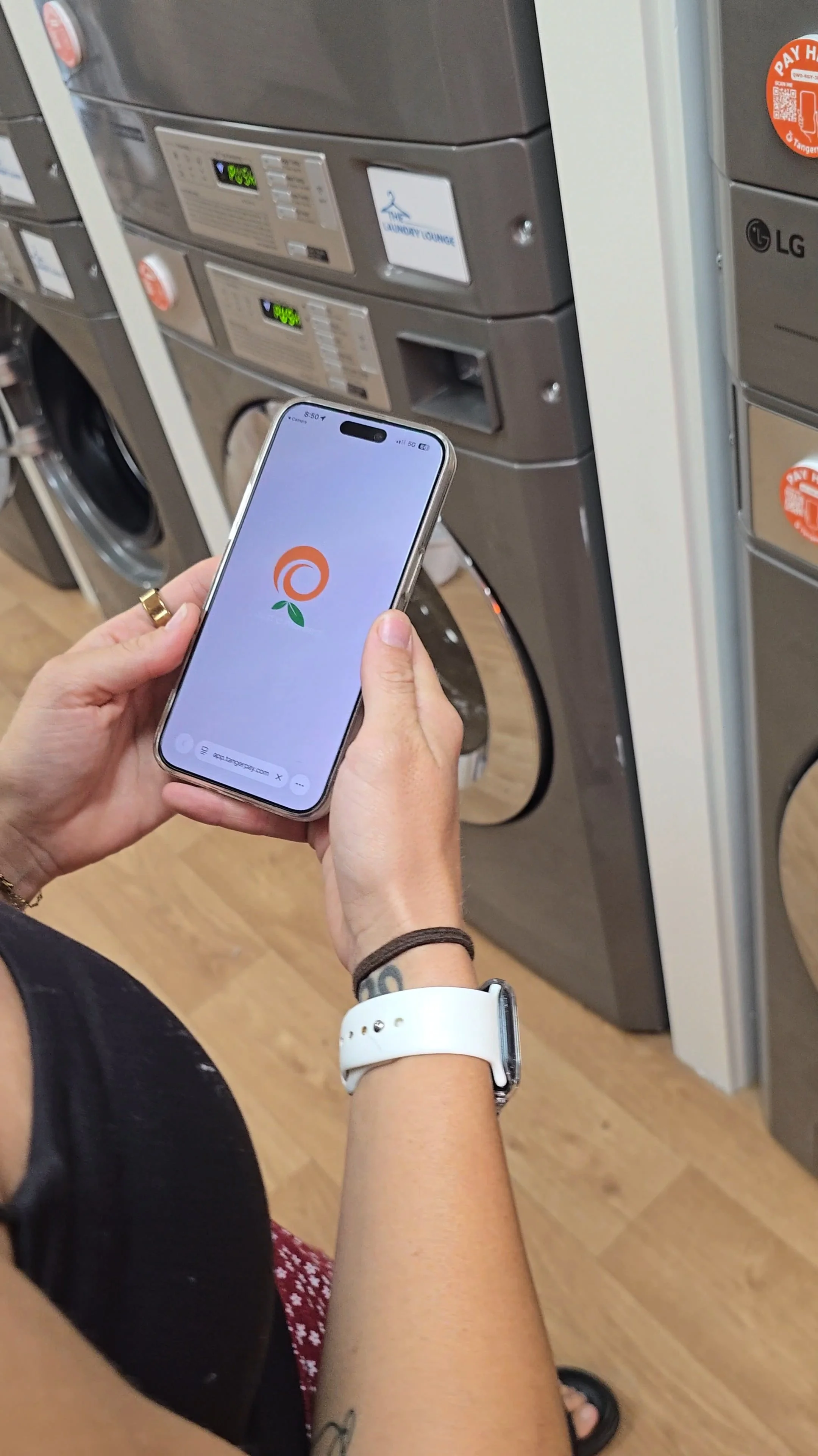 Person holding a smartphone with a logo on the screen, standing in front of a row of washing machines in a laundry room.