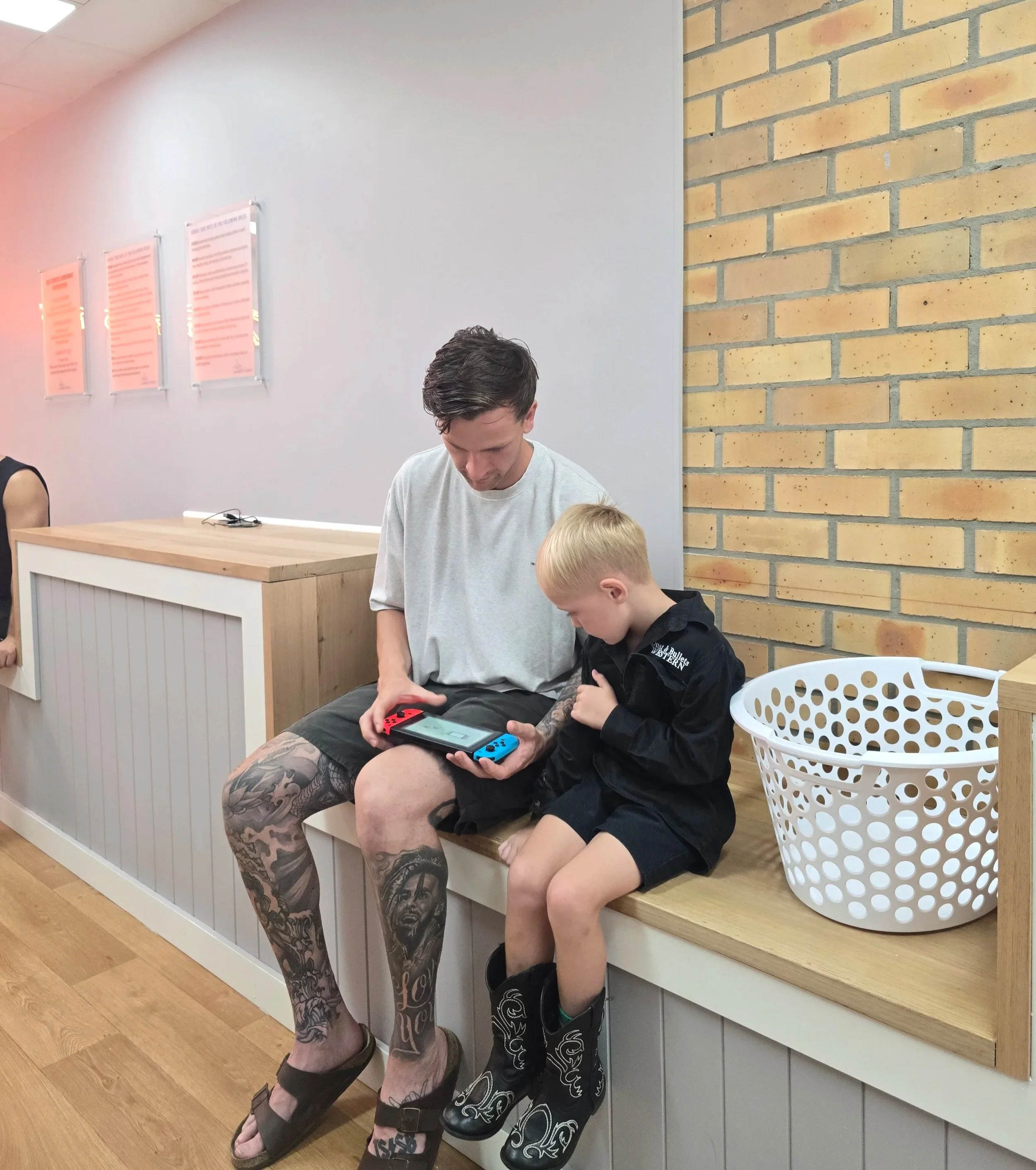 A man with tattoos on his legs and a young boy are sitting together on a wooden bench, focused on a handheld gaming device. There is a white laundry basket beside them, and the background includes a white wall with three pink framed notices and a brick wall.