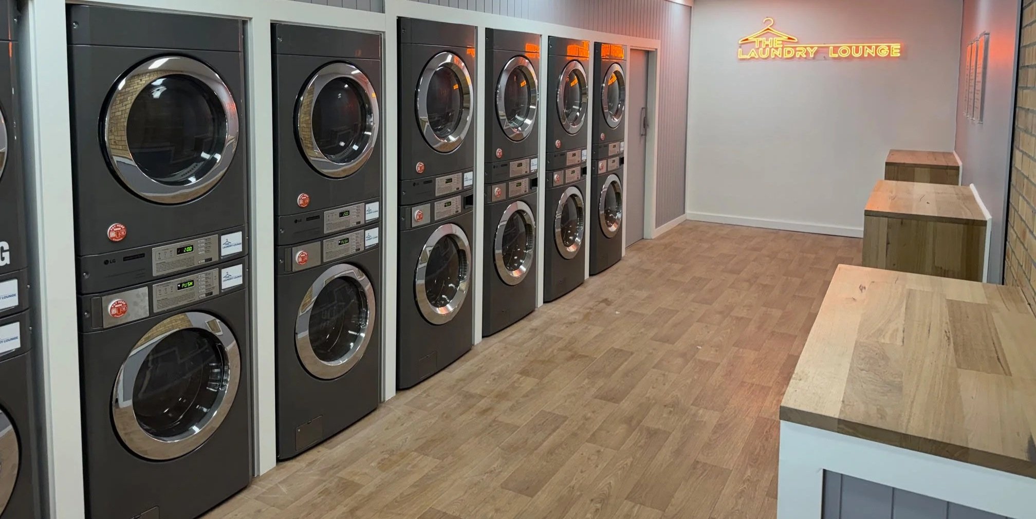 A laundromat with six front-loading washing machines lined up against a wall, a neon sign that reads 'The Laundry Lounge' on the wall, and wooden countertops along the right side.