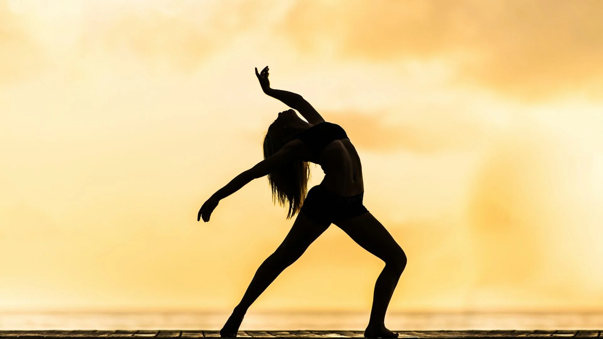 Silhouette d’une femme dans une posture de danse, bras levés et tête en arrière, sur un ciel nuageux jaune, illustrant la méditation Corps comme refuge pour débutants, pour habiter le corps et retrouver stabilité, calme et confiance.