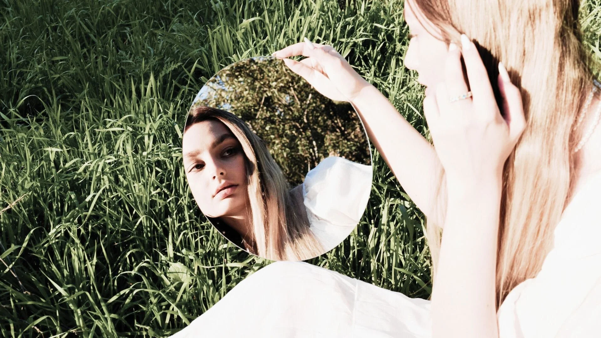 Femme blonde portant une robe blanche assise dans l’herbe, tenant un miroir rond où se reflète son visage, illustrant la méditation Jardin des miroirs, une visualisation guidée pour un voyage intérieur et une profonde réflexion sur soi.