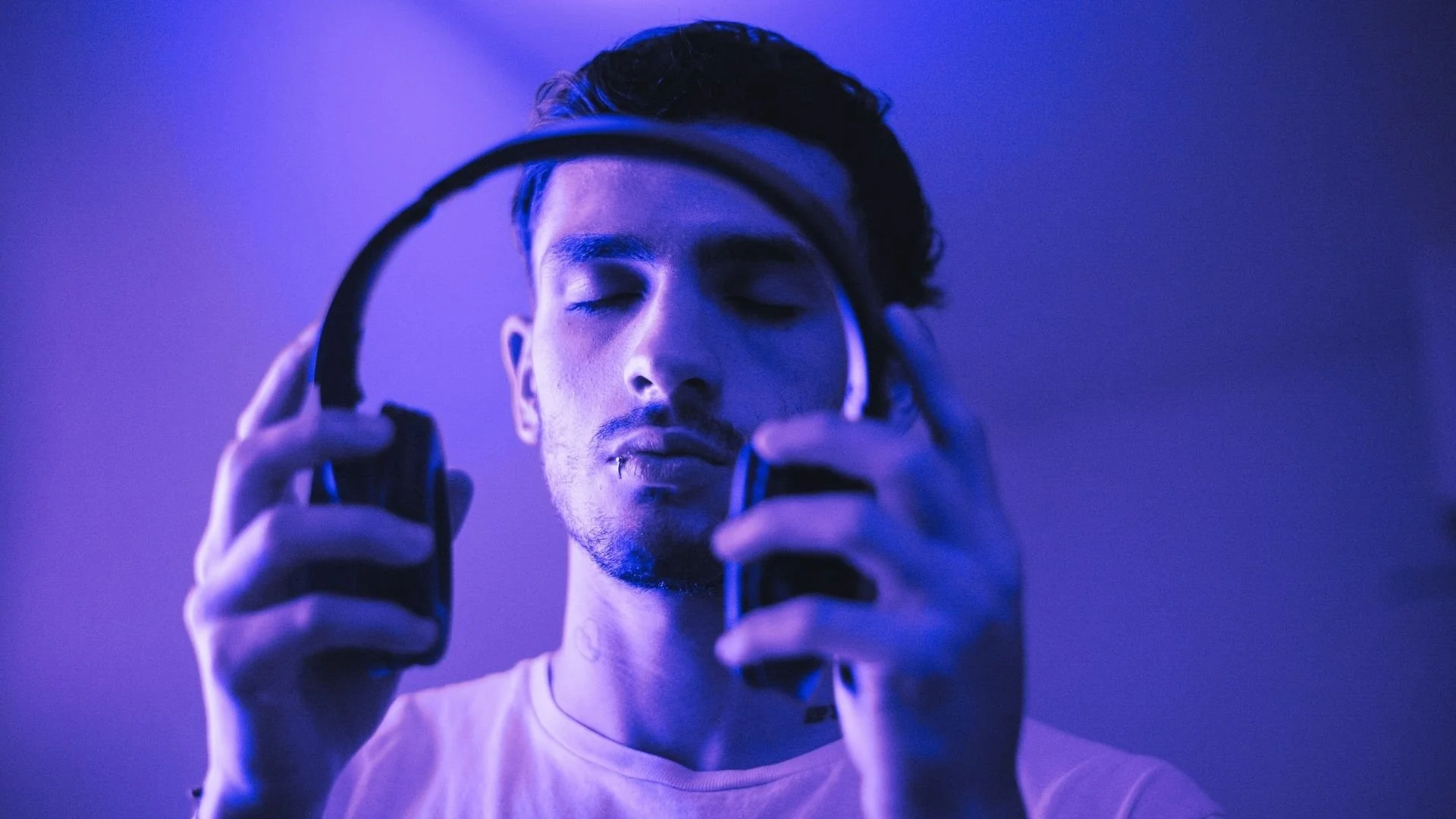 Homme adulte aux yeux fermés, portant une boucle d’oreille à la lèvre, sur le point de mettre ses écouteurs, sur une image légèrement bleutée, invitant à explorer la catégorie Zen Toi et sa variété de méditations pour tous les niveaux.