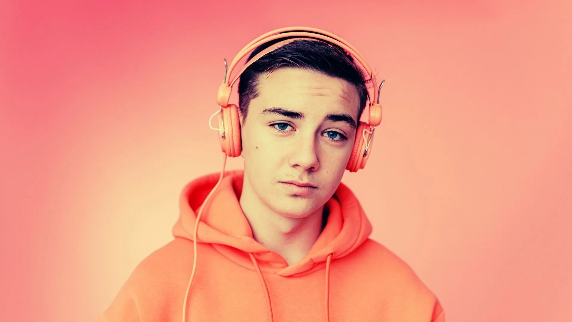 Adolescent au regard malicieux, chandail et écouteurs orange sur fond orangé, illustrant la catégorie Zen Attitude avec des méditations guidées pour adolescents.