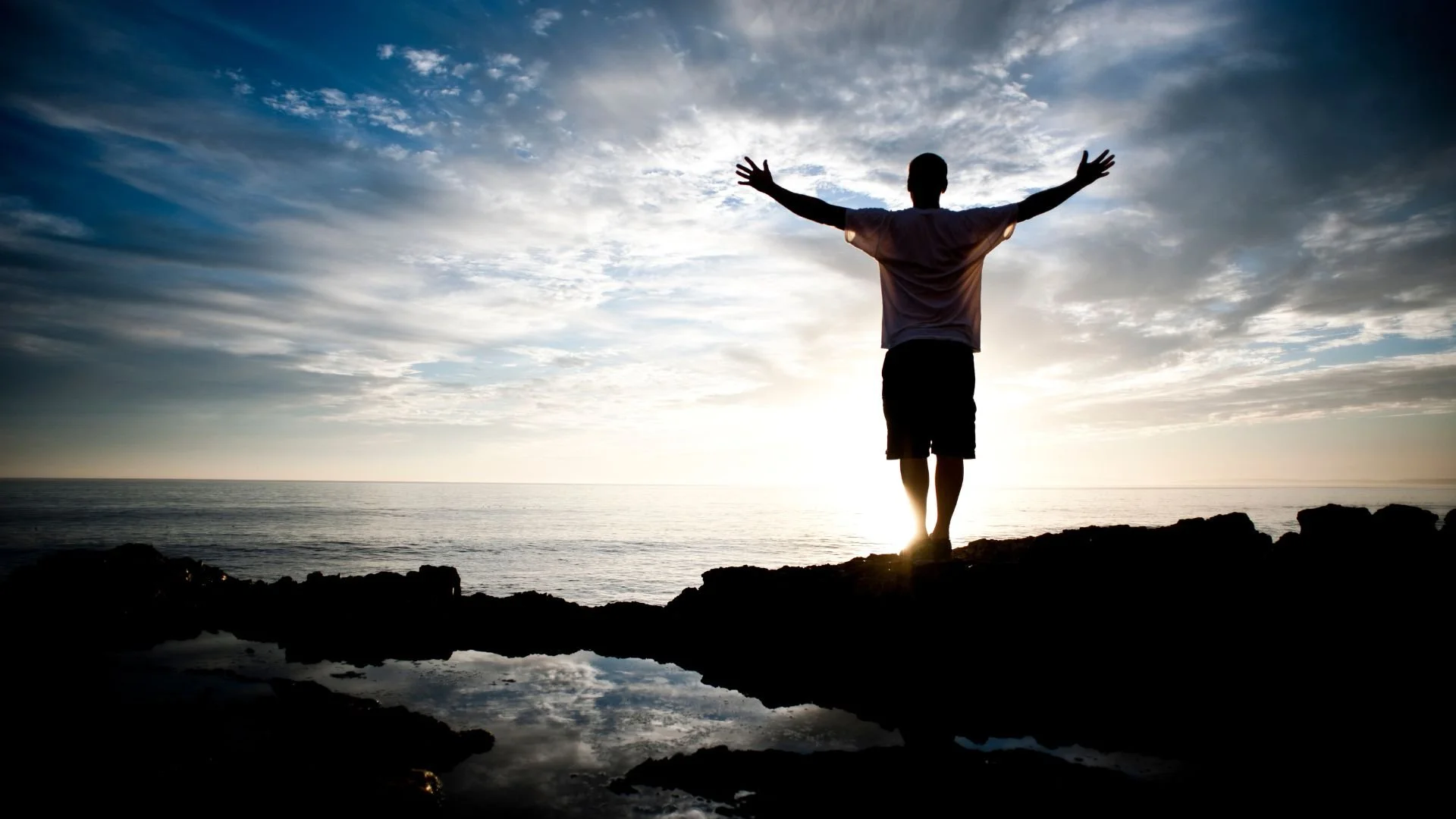 Homme debout sur un rocher face à l’océan à perte de vue, bras ouverts sous un ciel spectaculaire, illustrant une méditation de gratitude pour se recentrer dans l’instant présent.