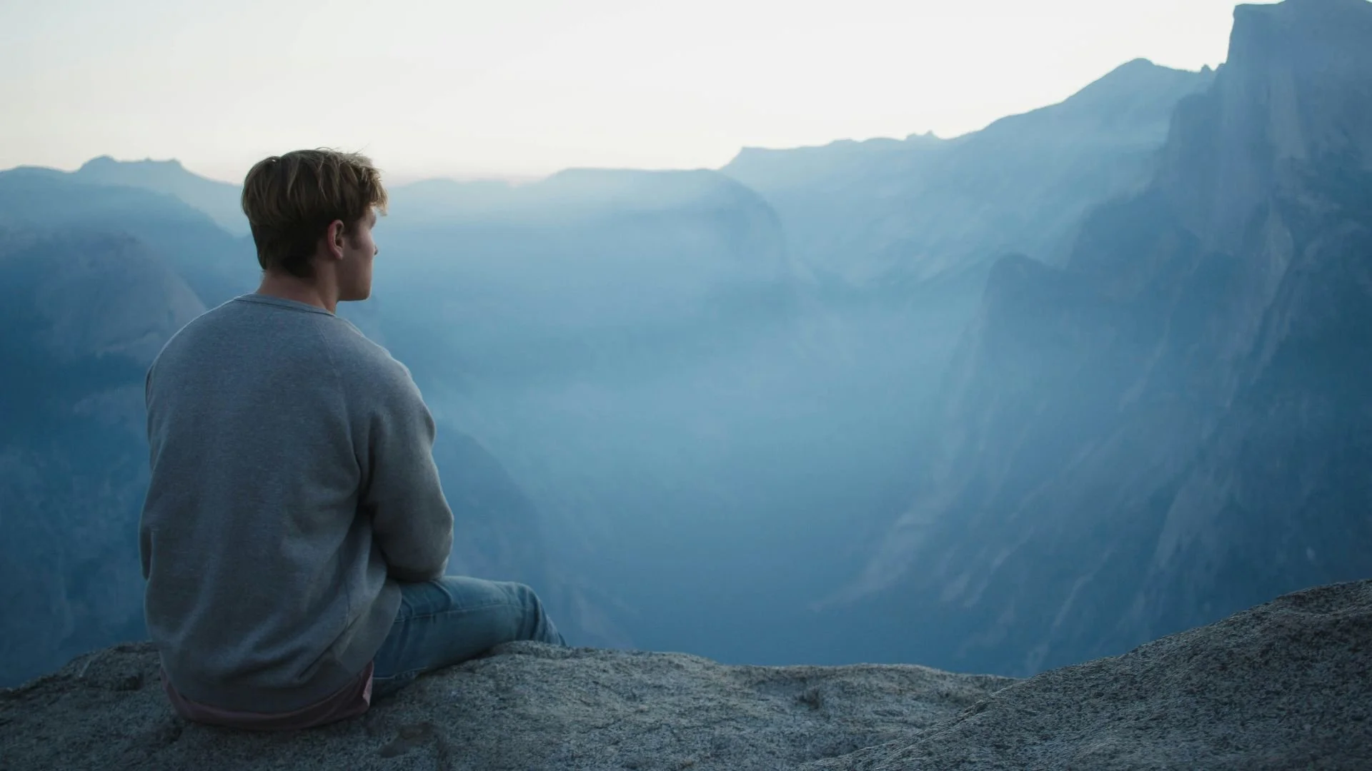 Jeune homme en jeans et sandales gris assis sur un rocher, regardant un paysage de montagne brumeux, illustration aux teintes bleu-gris pour la méditation Espace intérieur de la catégorie Zenitude 101, pour créer de l’espace et se sentir posé.