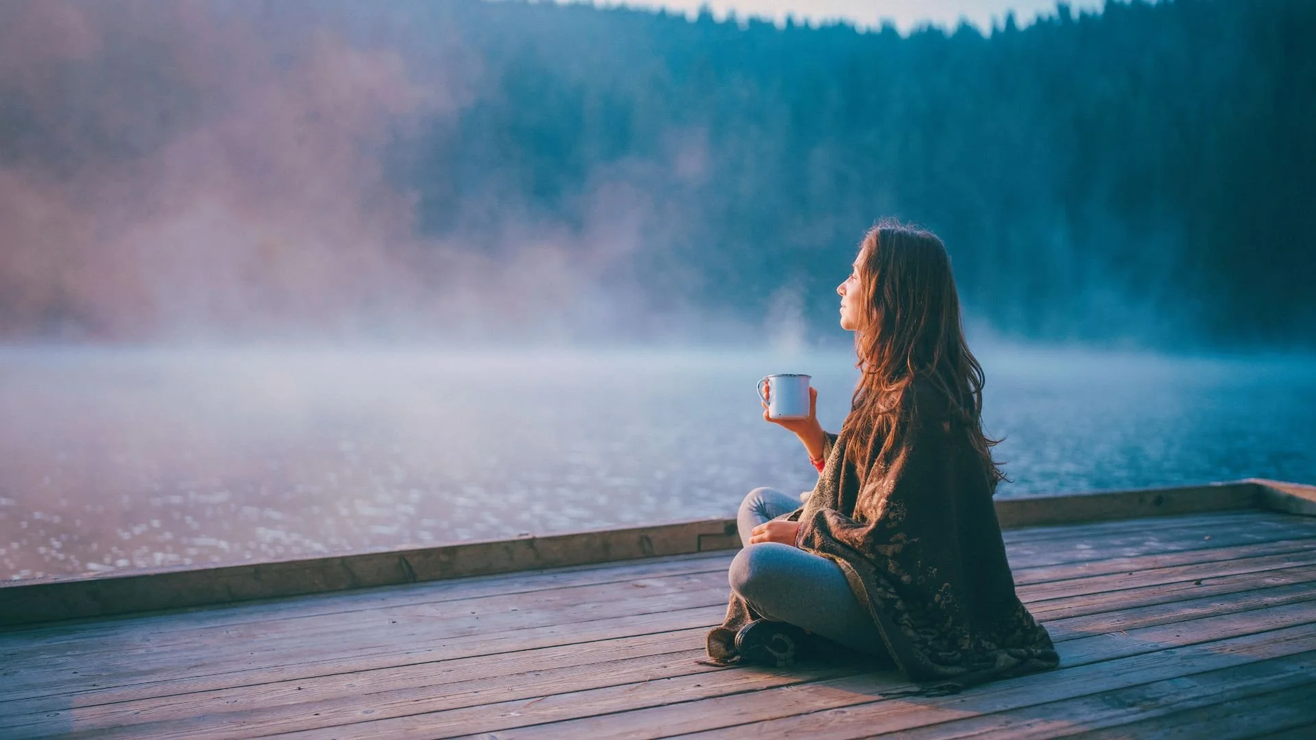 Femme assise en indien sur un quai devant un lac brumeux, tenant une tasse, image bleutée et rosée, illustrant la méditation Détente profonde pour relâcher les tensions et se reconnecter à une sensation de sérénité.