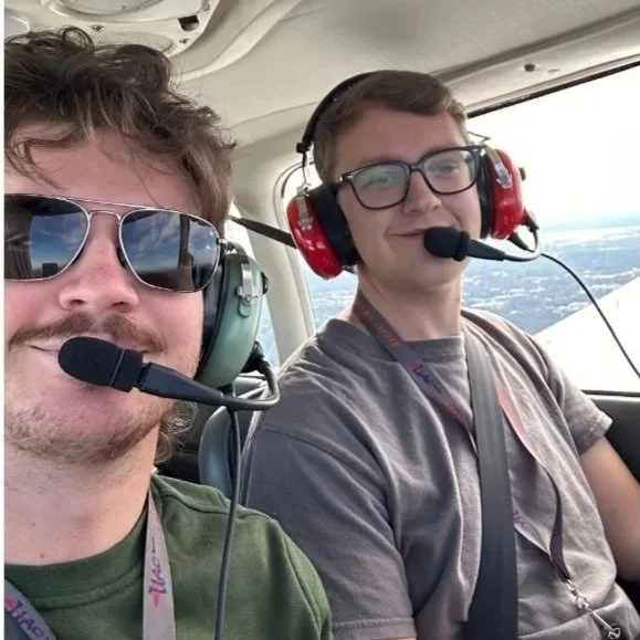 Two men sitting in a helicopter cockpit wearing headsets with microphones, with a view of the sky and landscape outside the window.