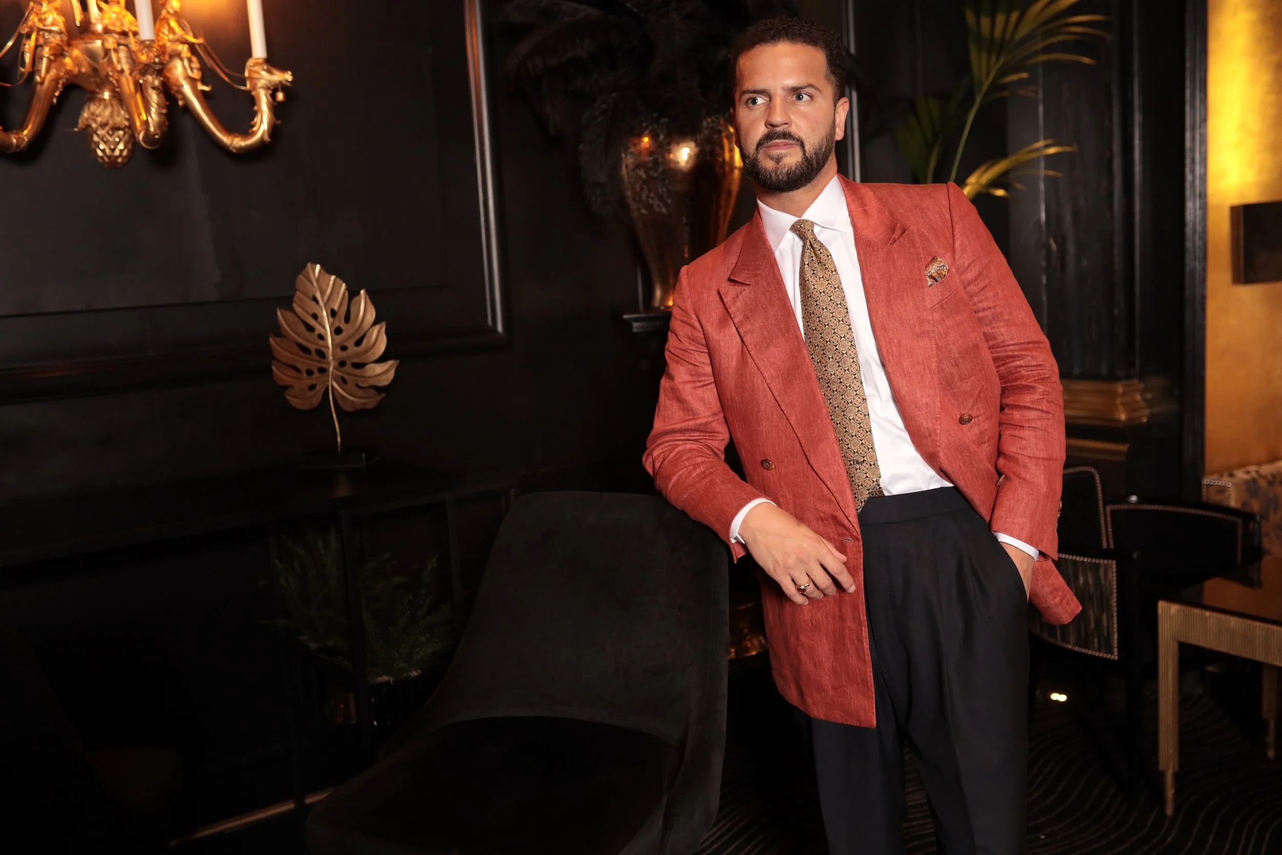 Man in a reddish-orange bespoke loro piana linen suit jacket, white shirt, and patterned tie, standing in a dimly lit, stylish room with black walls, gold decor, and plants.