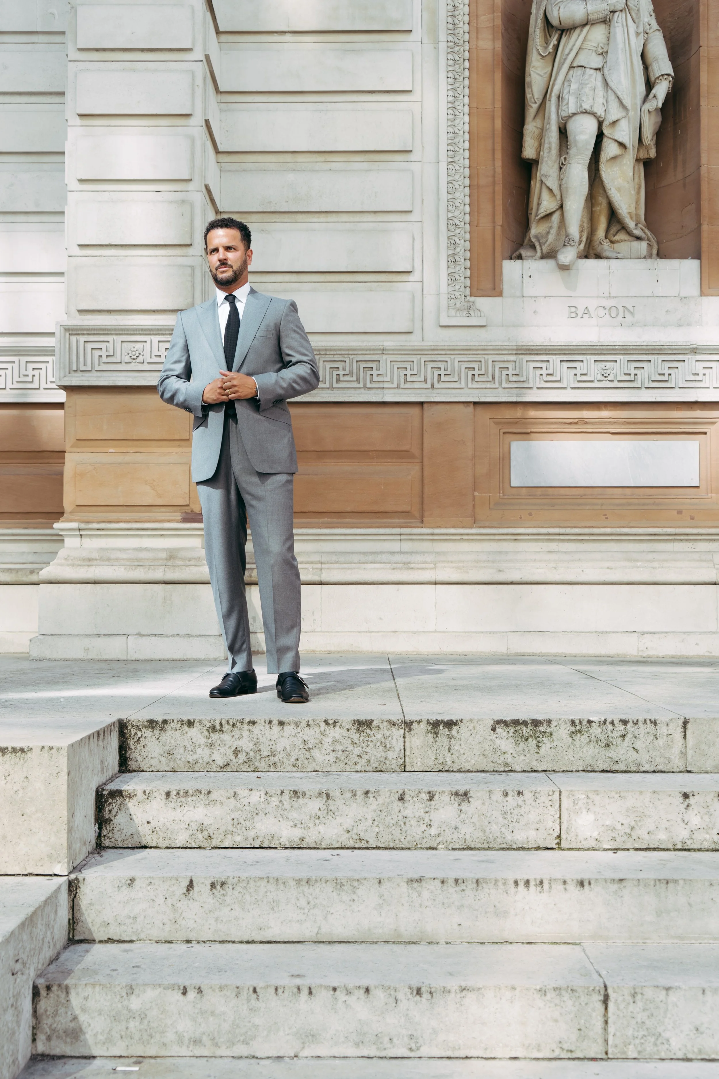 A man in a grey wool bespoke suit standing on the steps outside a building with classical architectural details and a marble statue in Mayfair, London.