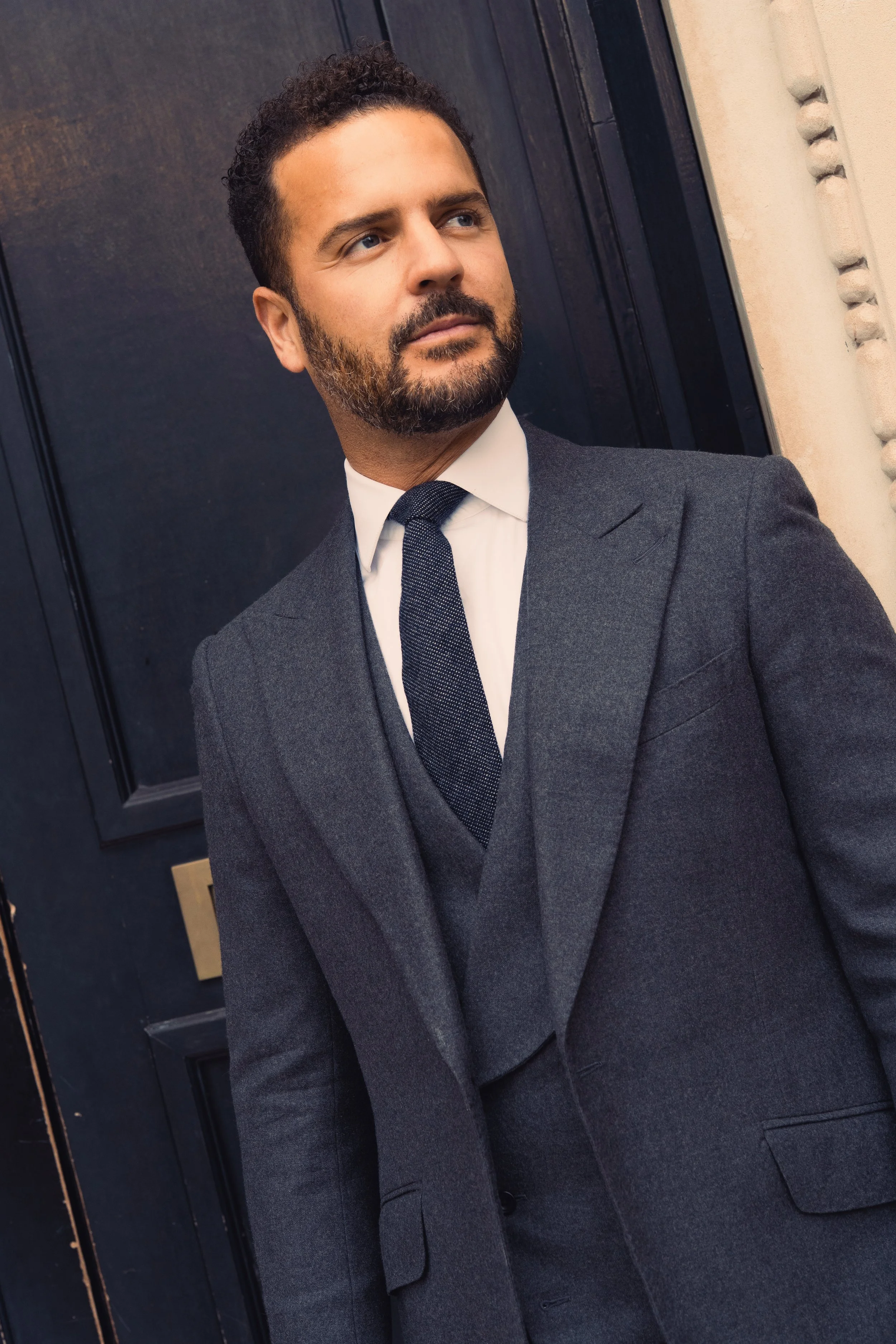A man in a dark grey flannel bespoke suit, cloth by Dormeuil, white shirt, and dark tie standing outdoors in front of a dark door and cream-colored wall.