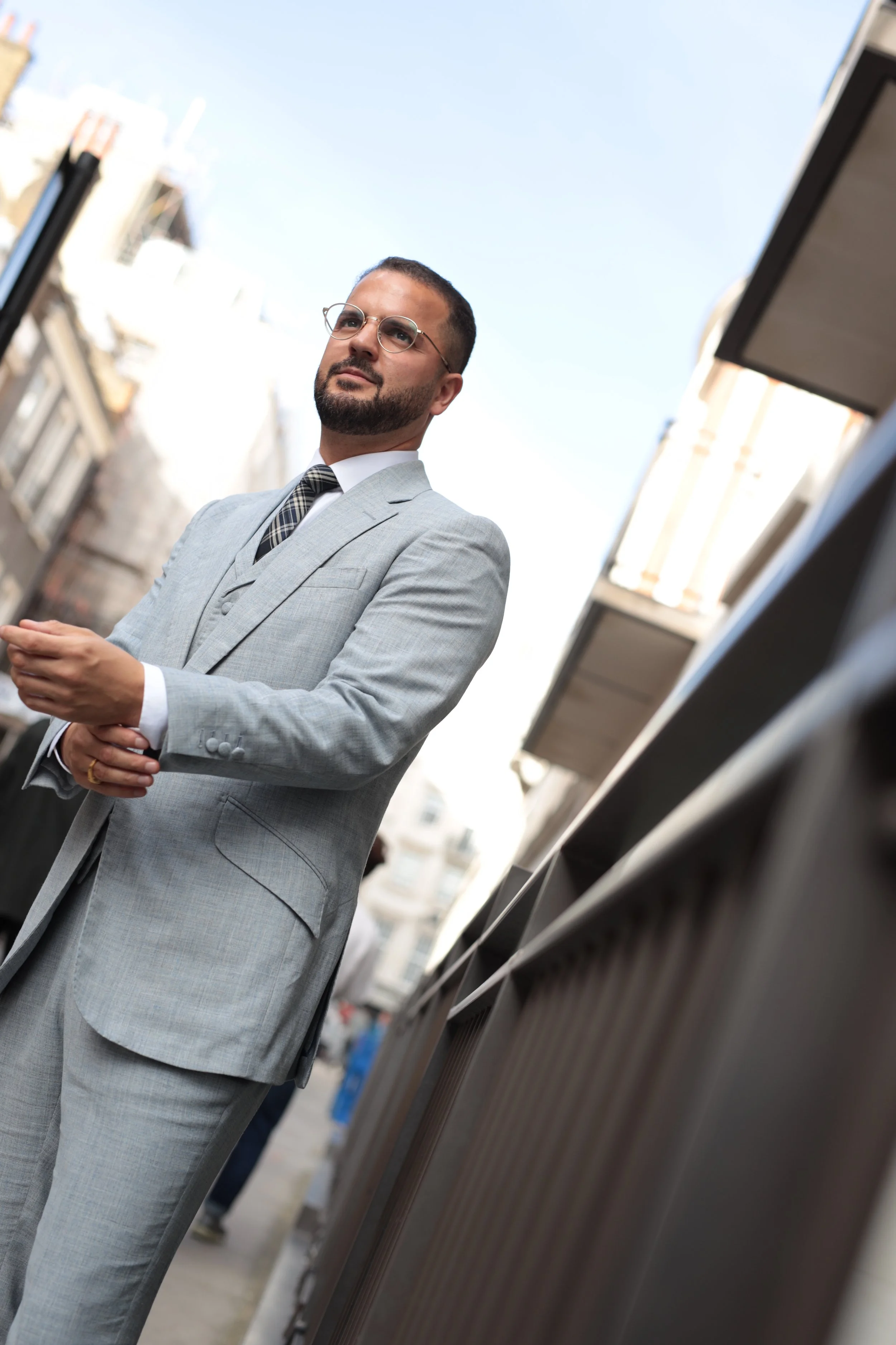 A man in a light grey bespoke, tailored suit, white shirt, and plaid tie stands outdoors in an urban area in London, looking thoughtfully into the distance with buildings and a clear sky in the background.