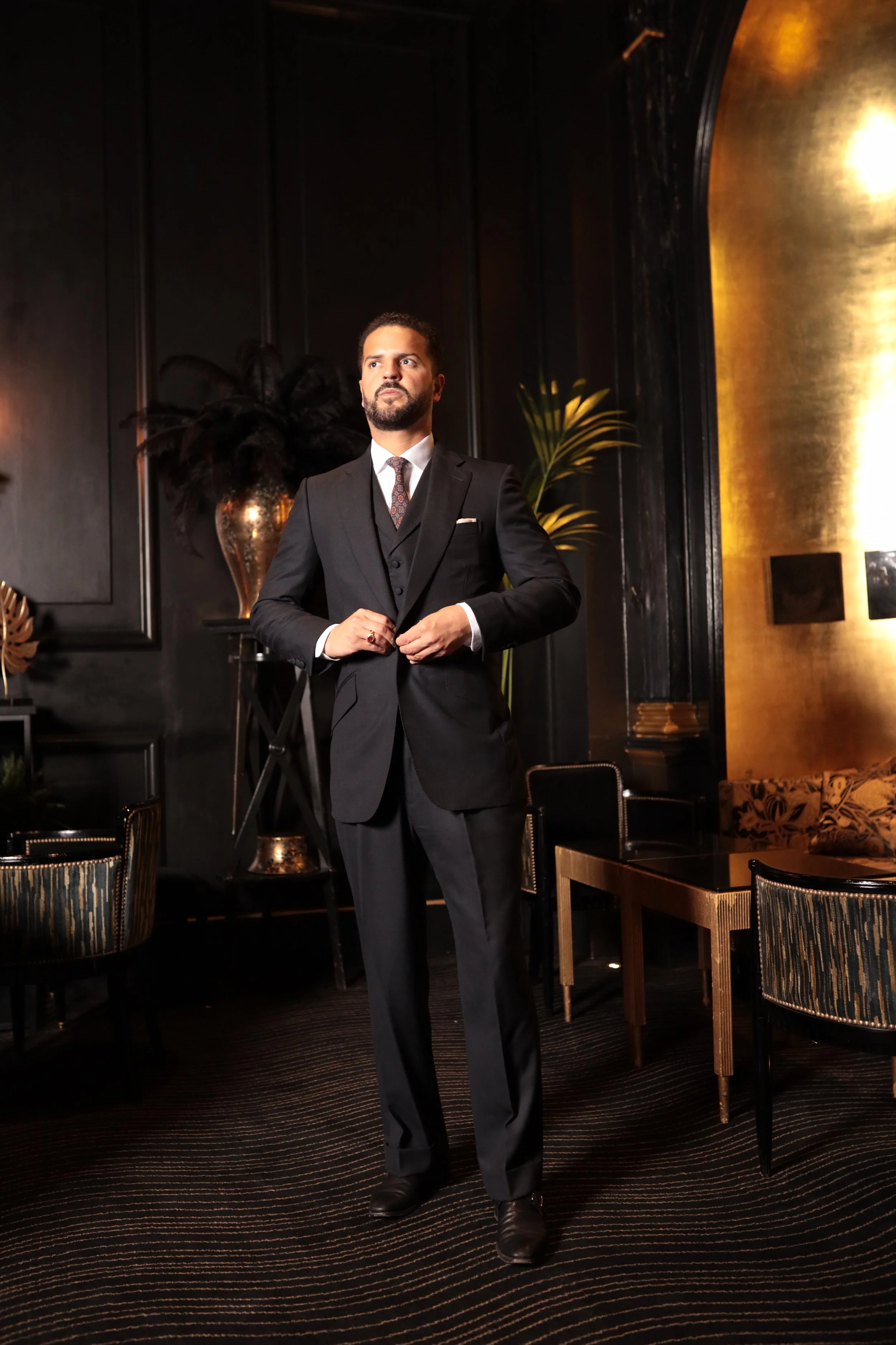 Man in a bespoke tailored navy suit standing in a luxurious, dimly lit room with dark walls, gold accents, and modern furniture.
