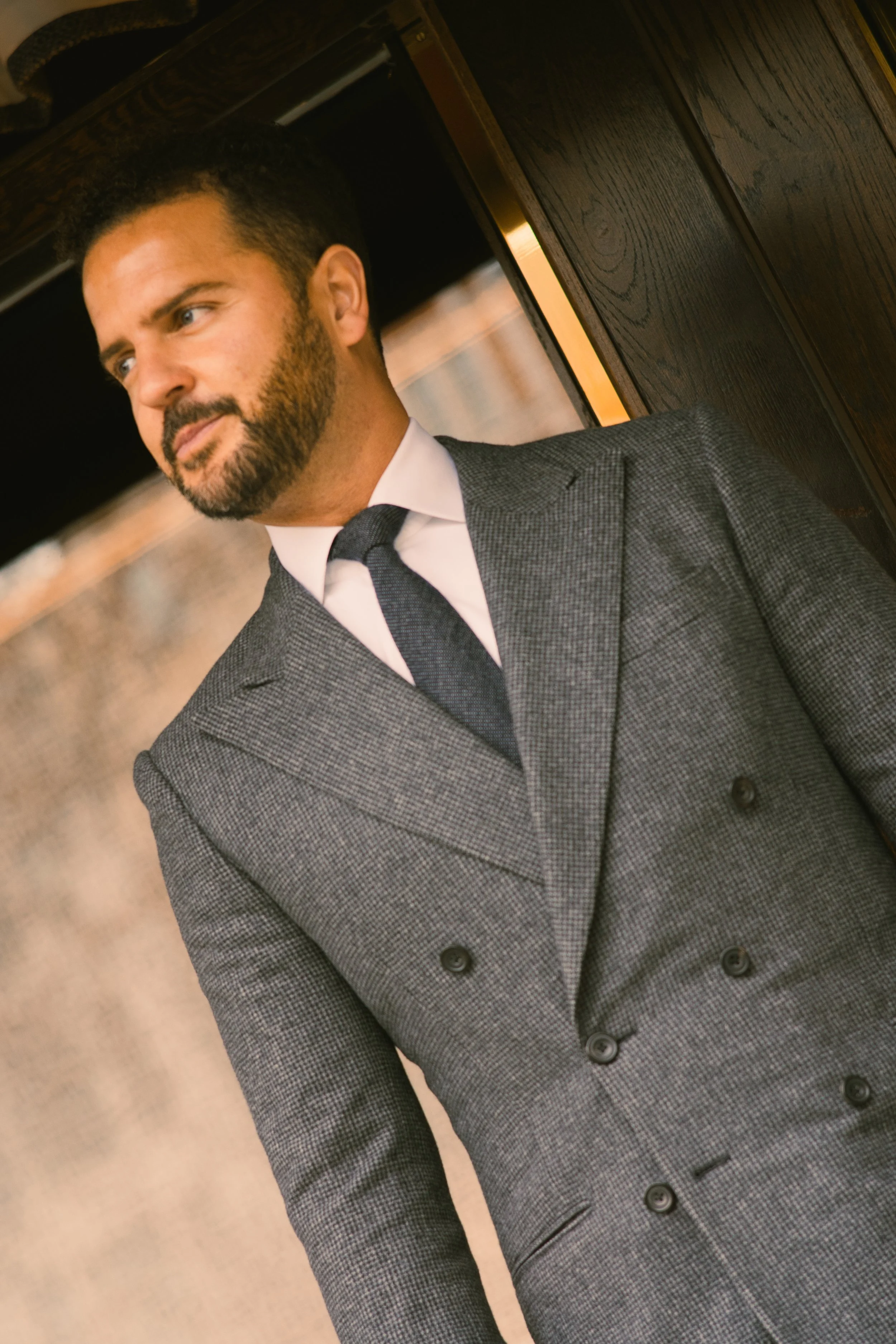 A man in a grey double-breasted flannel suit, white cotton dress shirt and black tie standing indoors near a wooden wall, looking thoughtfully to the side.