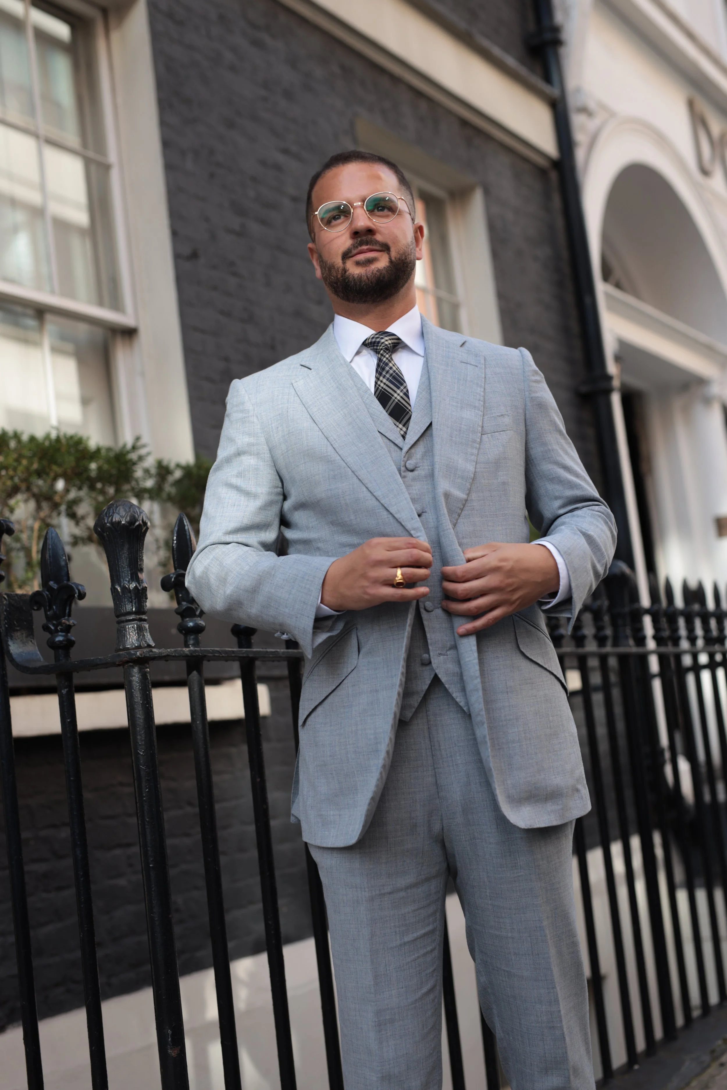 A man in a loro piana light grey and blue bespoke summer suit standing in front of a black wrought-iron fence and a dark gray building, adjusting his suit jacket, wearing glasses and a patterned tie.