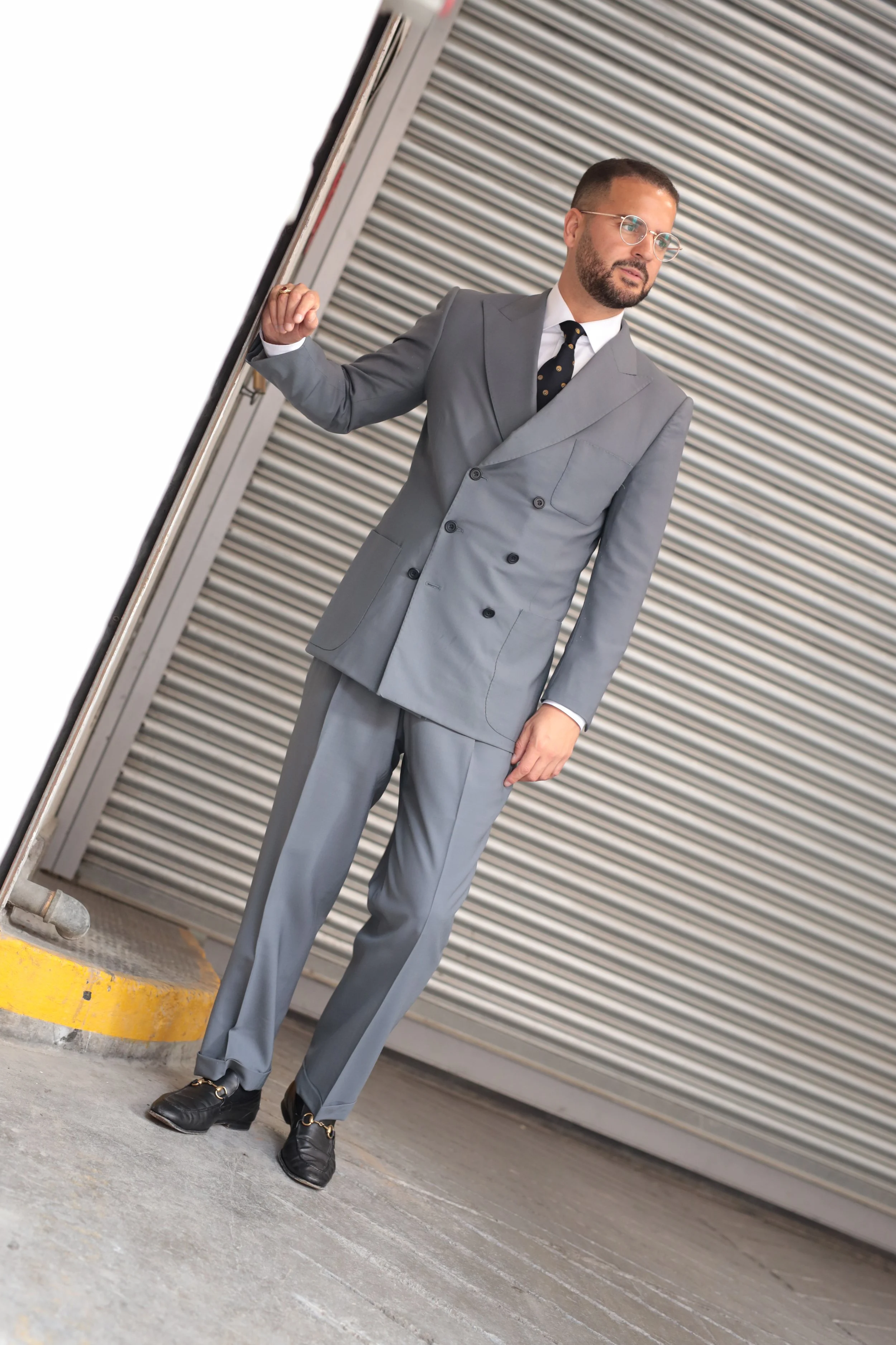 A man wearing a tailored grey double-breasted suit, black gucci shoes, and glasses standing next to a partially open roller shutter door in an urban setting.
