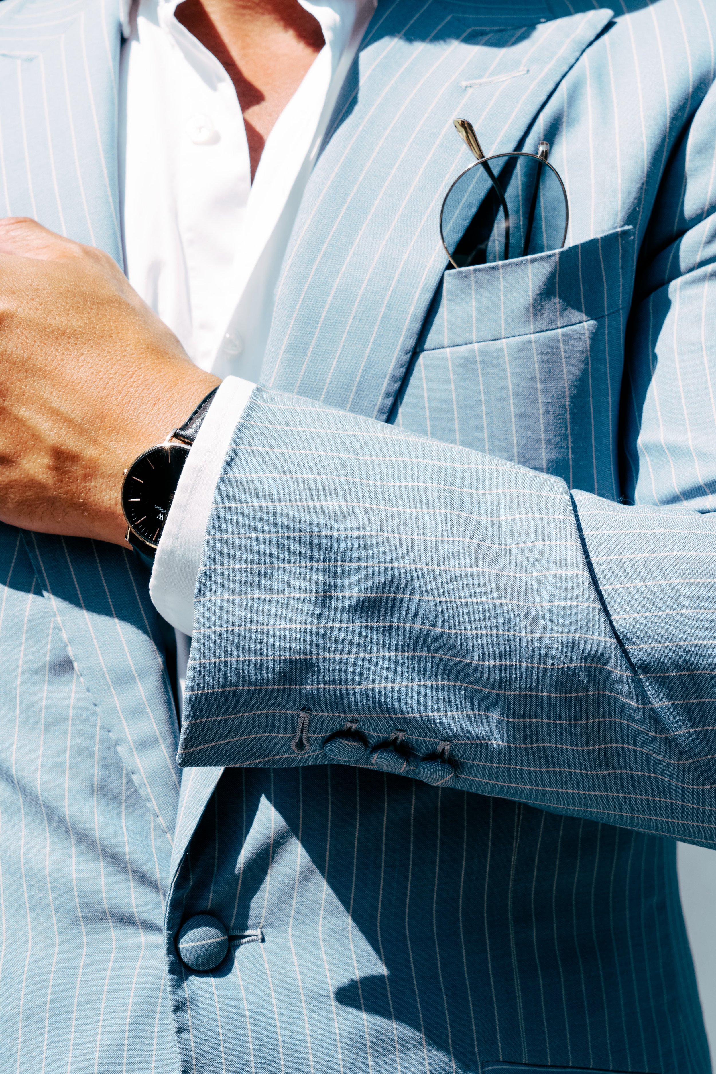 Close-up of a man wearing a light blue bespoke tailored pinstripe suit with a white shirt, a wristwatch, and sunglasses in the suit pocket, with his arms crossed.