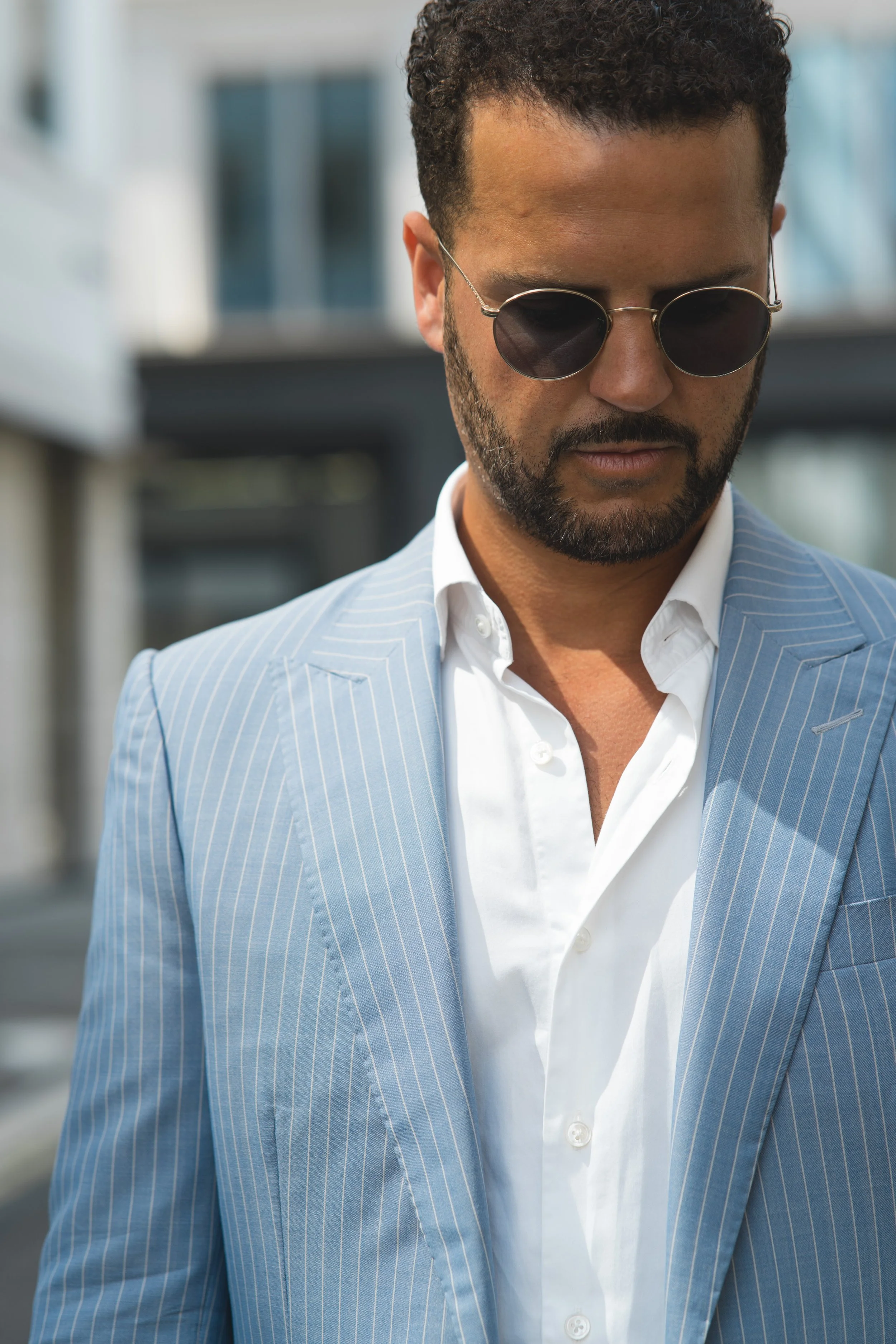 A man wearing sunglasses, a light blue tailored pinstripe suit, and a white shirt, looking down outdoors in London.