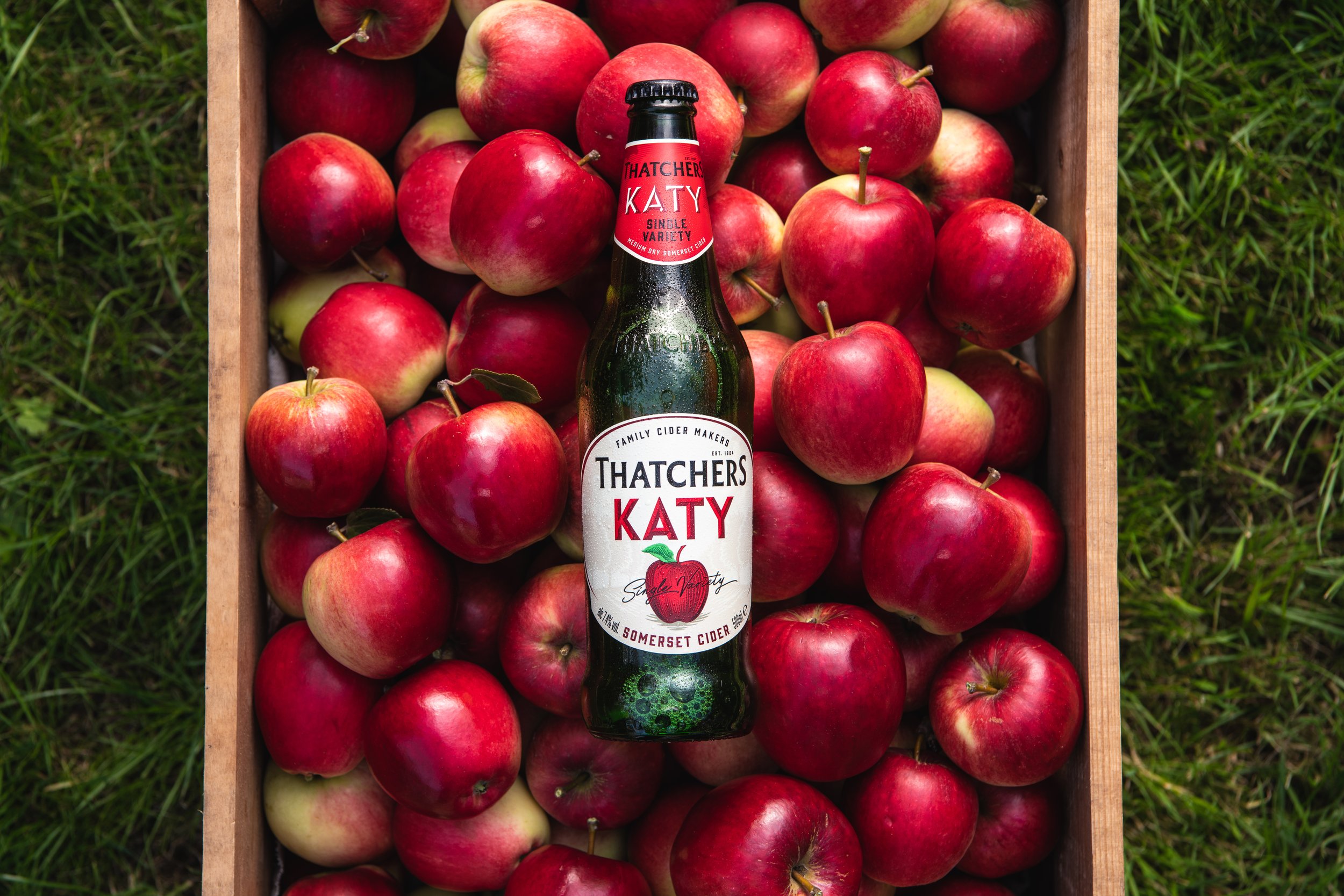 Láhev cideru Thatchers Katy stojí na dřevěné bedýnce plné červených a zelených jablek, položené v zelené trávě – čerstvá, letní atmosféra jako dělaná na chill i piknik.