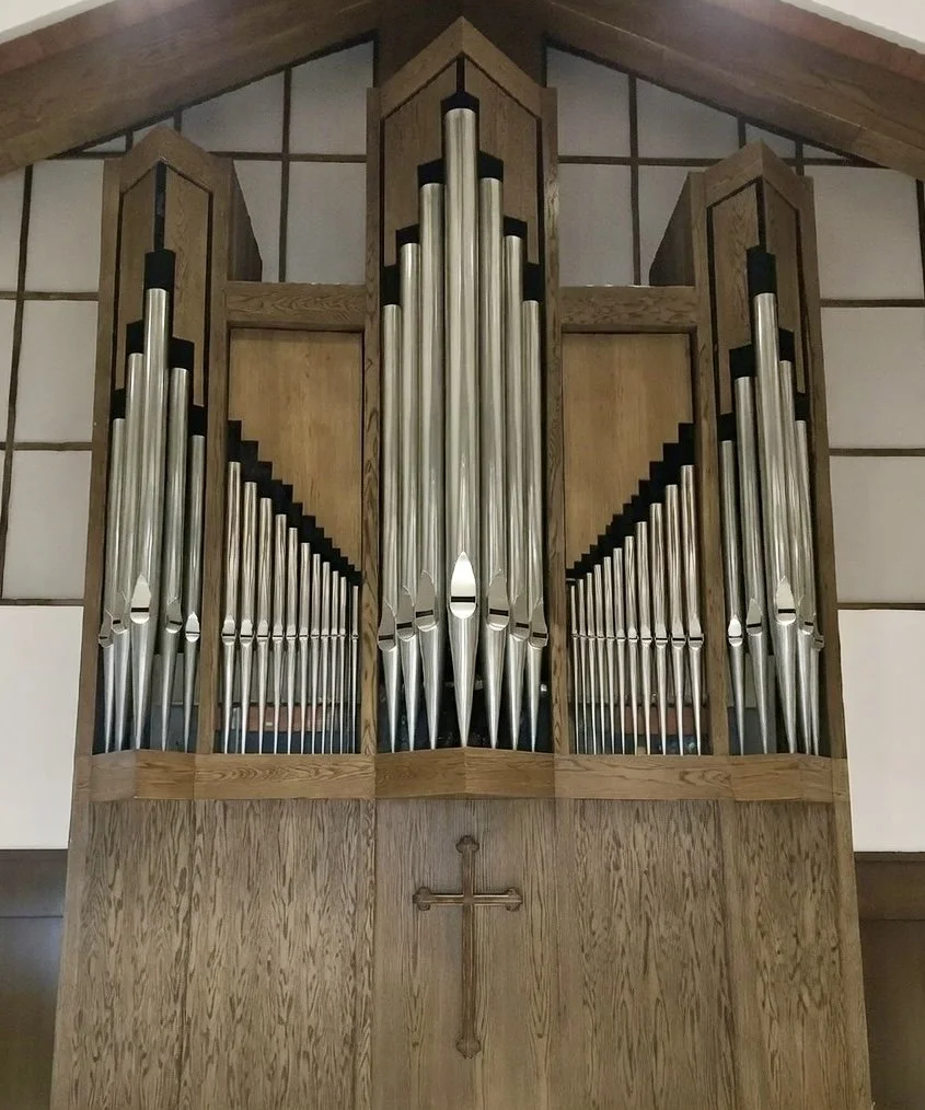 A church pipe organ with metallic pipes in a wooden case, featuring a cross symbol on the wood panel below the pipes.